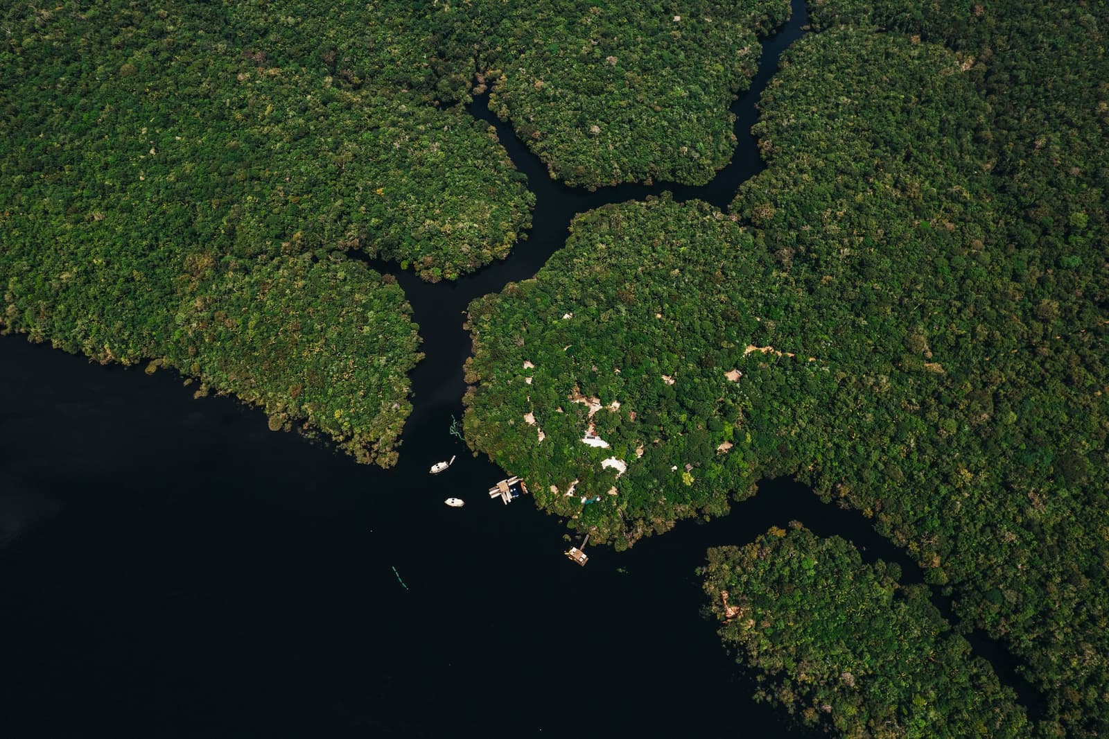 Tiny boats, vast forest, the lodge finds its place between the two. Aerial view of Anavilhanas Jungle Lodge Brazil, with small boats and a floating dock nestled at the edge of a dense rainforest island amid dark Rio Negro waterways