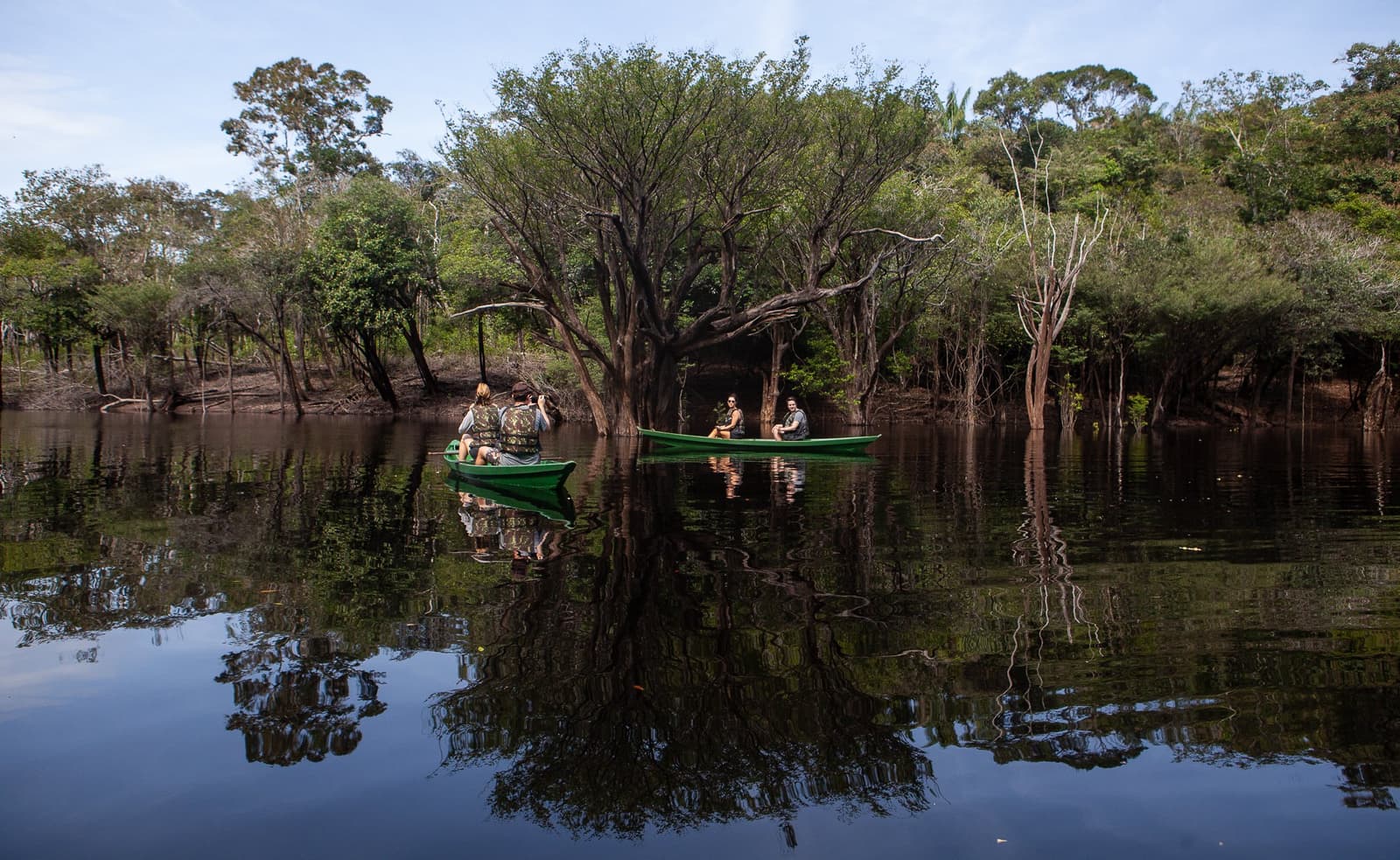 The river gives you two forests for the price of one. Three travellers in a green canoe gliding through a flooded forest on dark mirror-still water at Anavilhanas Jungle Lodge Brazil, surrounded by partially submerged trees and their perfect reflections