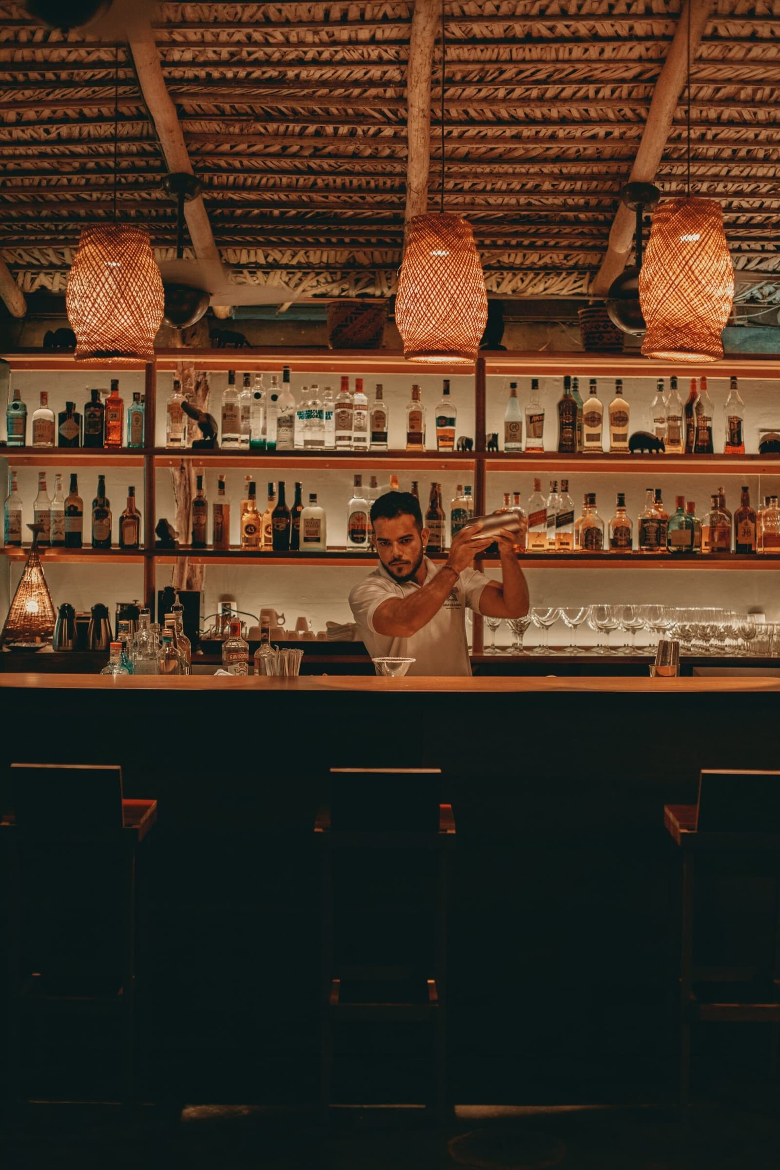 The jungle day winds down, and someone's already on it. A bartender shaking a cocktail shaker behind the bar at Anavilhanas Jungle Lodge Brazil, beneath warm woven rattan pendant lights and shelves lined with spirits