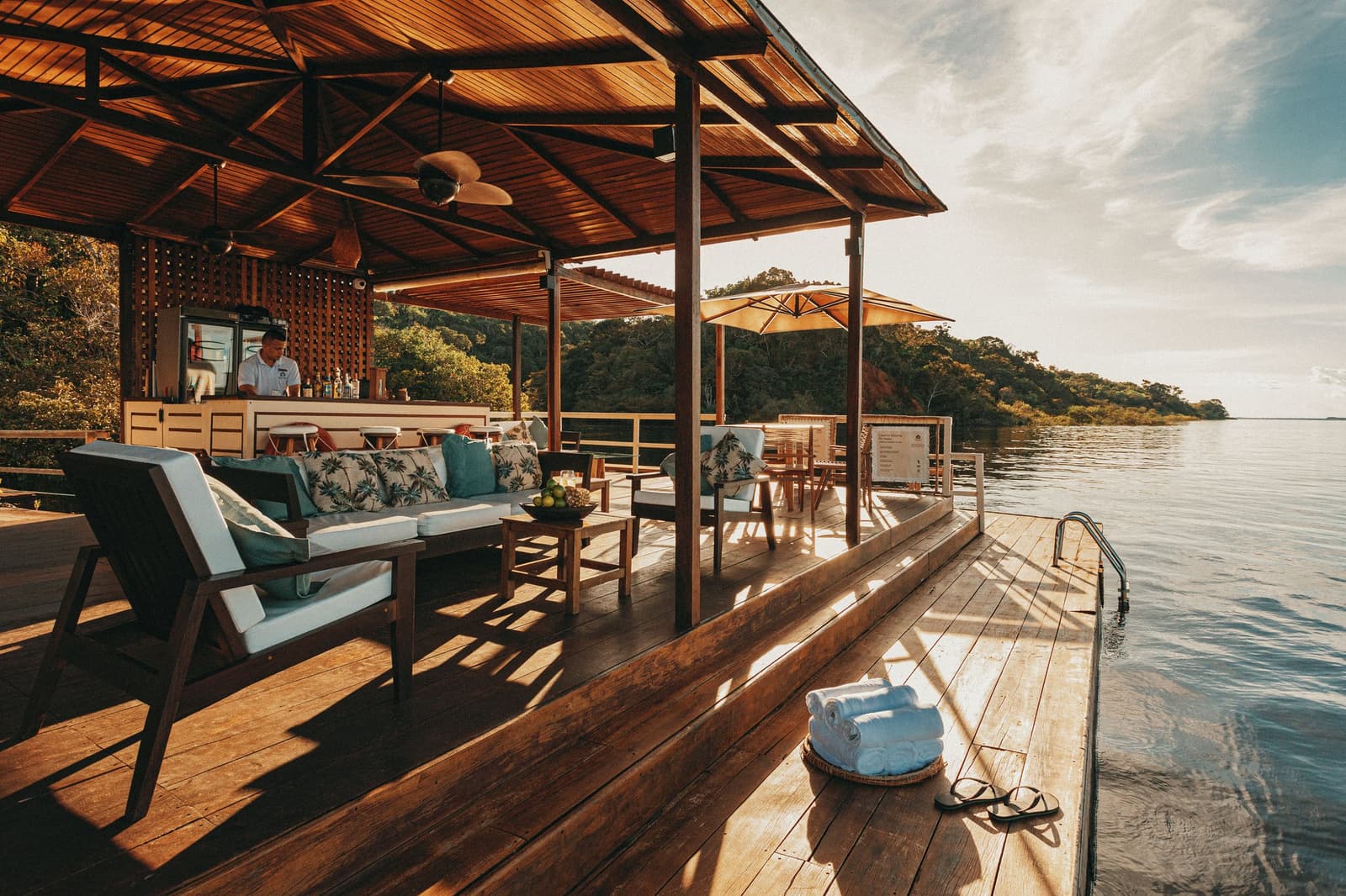 Sundowners taste better when the Amazon is your swimming pool. A warm-toned hardwood deck at Anavilhanas Jungle Lodge Brazil extends over the calm Rio Negro, with cushioned lounge seating, a staffed bar, folded towels by the water's edge, and forested islands in the background