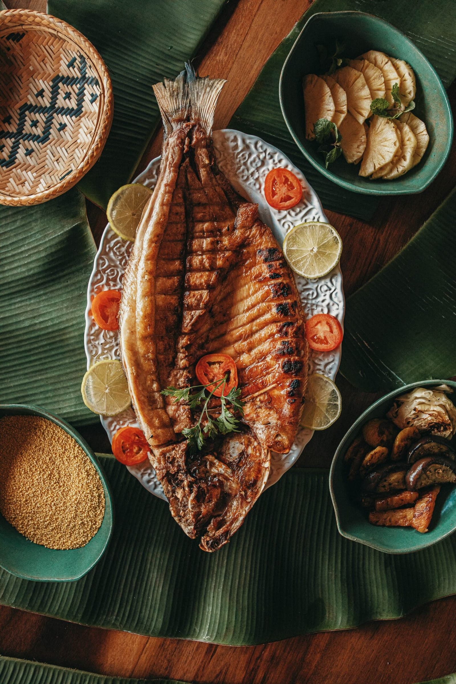 River-to-table, the way the Amazon has always done it. A whole grilled fish served on a decorative white platter with lime and tomato slices at Anavilhanas Jungle Lodge Brazil, surrounded by side bowls of pineapple, roasted mushrooms and golden seeds on banana leaves