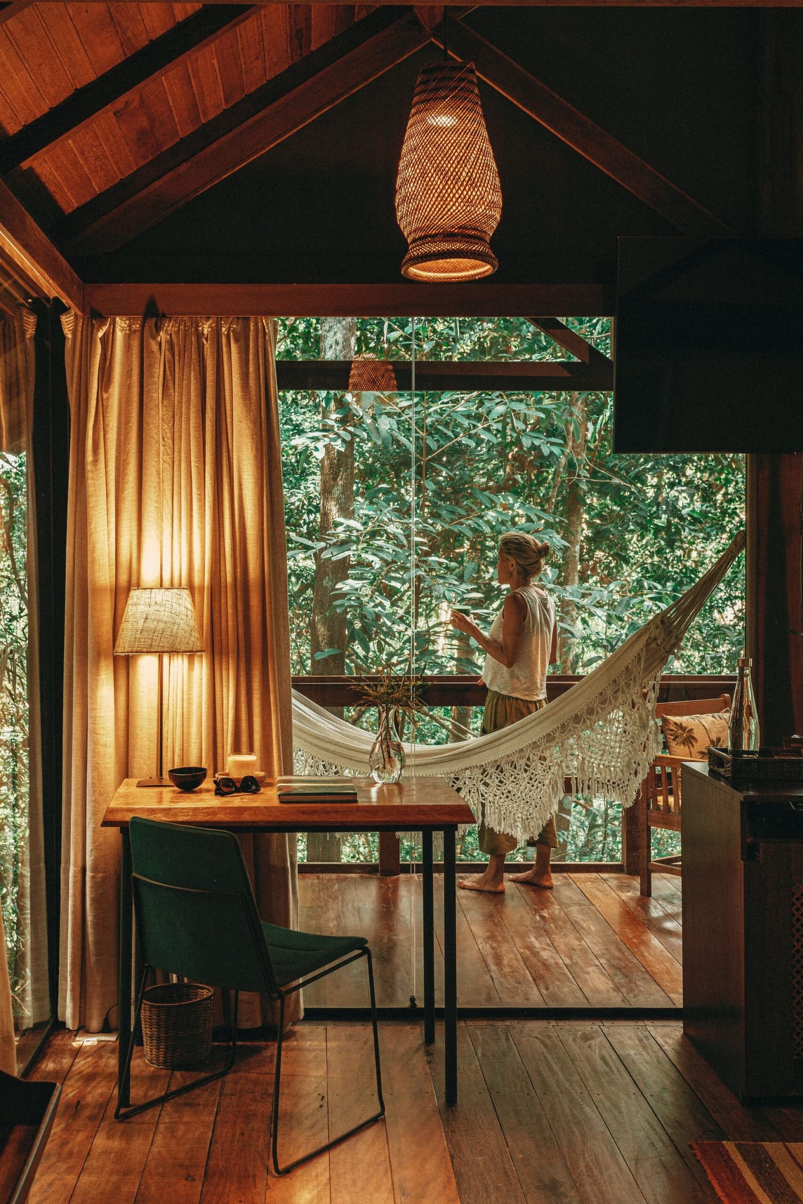 Coffee, forest, hammock. Some mornings sort themselves out. Interior of a luxury Amazon lodge cabin with warm timber floors, a woven pendant lamp, and a woman standing barefoot on a hammock-hung veranda gazing into dense green rainforest