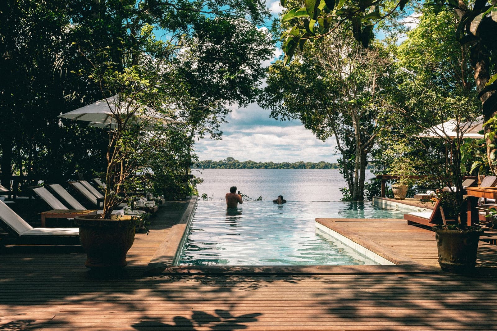 The pool ends where the river begins, that's the whole idea. Two guests floating in the infinity pool at Anavilhanas Jungle Lodge Brazil, framed by dense rainforest trees with the wide Rio Negro stretching to the horizon
