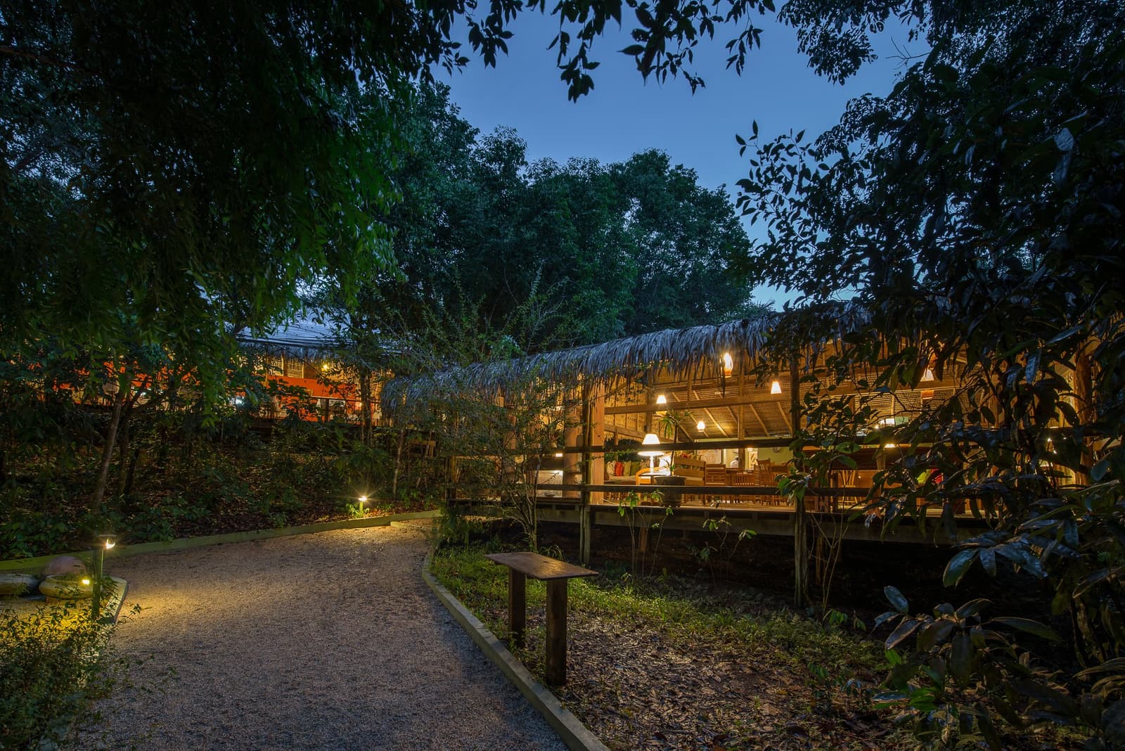 The lodge calls you in at dusk, warm lights threading through the forest. A gravel pathway lined with low bollard lights leads through dense tropical trees toward the open-sided thatched dining pavilion of Anavilhanas Jungle Lodge Brazil, glowing warmly at dusk beneath a deep blue twilight sky