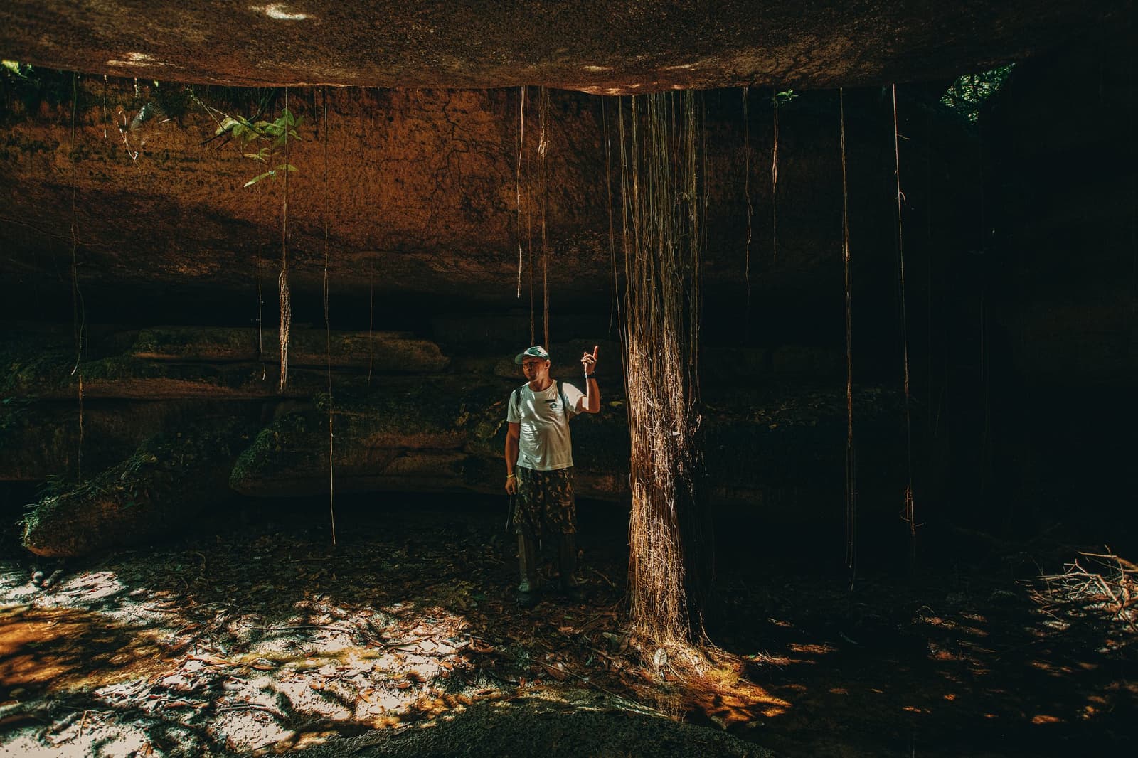 Your guide reads the forest like a book, even underground. A guide points upward beneath a massive ancient rock overhang at the Madadá Caves, Anavilhanas Jungle Lodge Brazil, with long aerial roots cascading from the ceiling in dramatic golden light