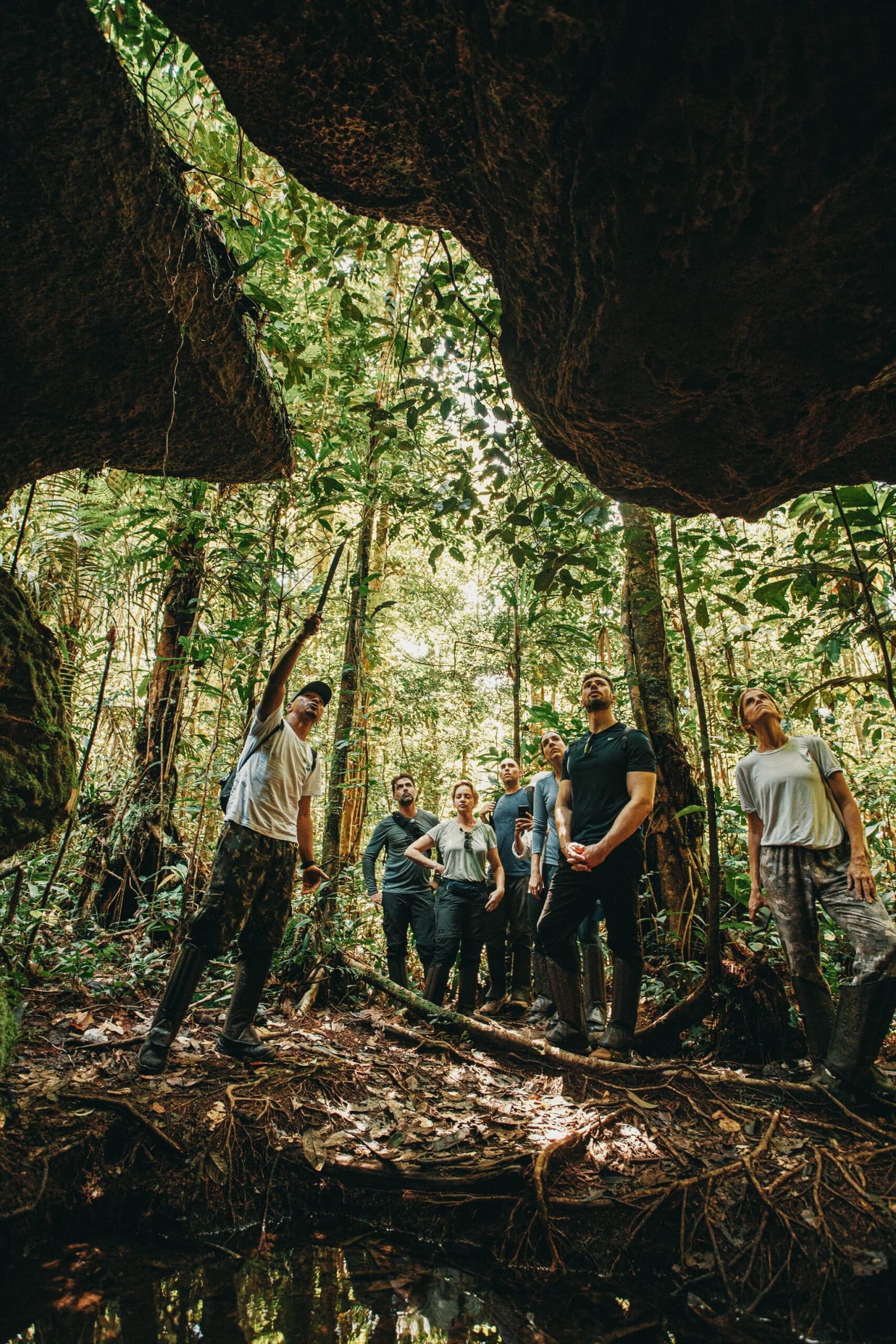 The forest has a way of making you look up and go quiet. A guide points upward while a small group of travellers gazes toward the rainforest canopy, framed by the ancient rock arch of a cave at Anavilhanas Jungle Lodge Brazil