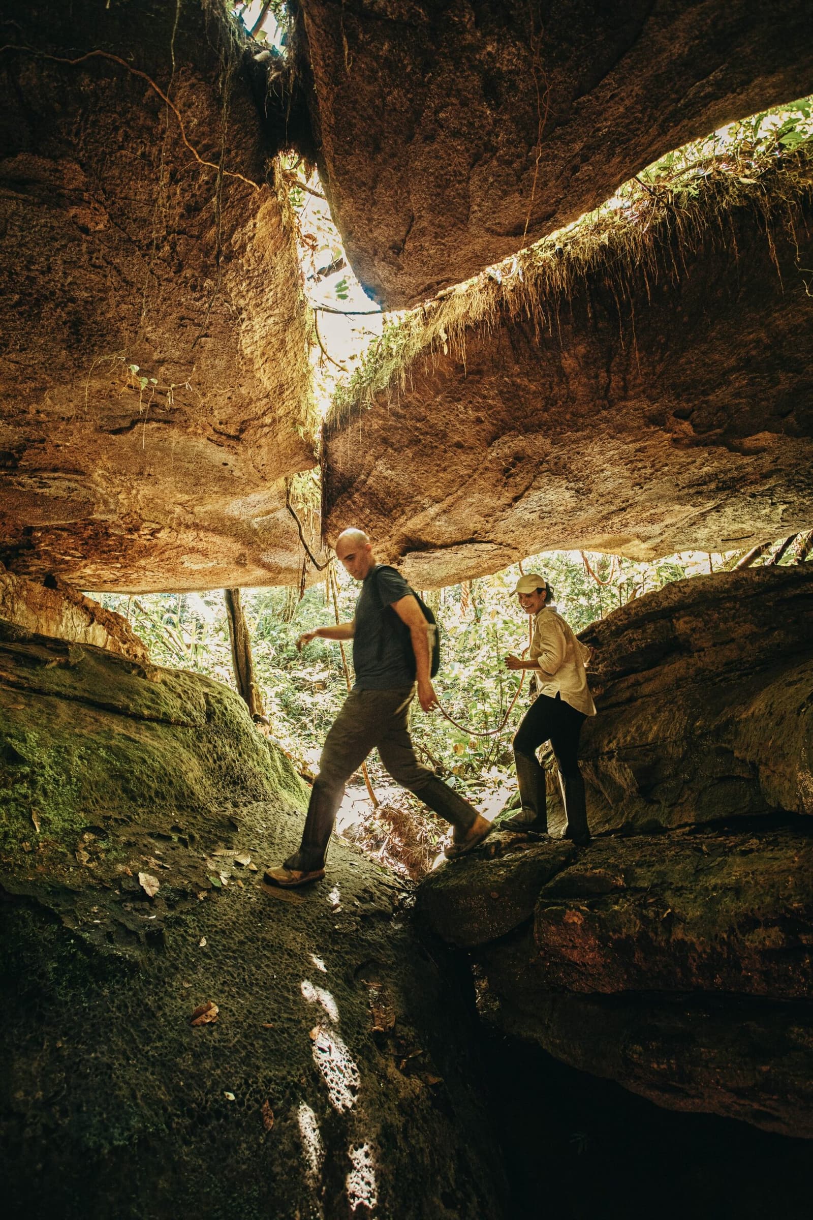 Some trails ask you to duck, step, and trust your footing a little. Two hikers duck beneath a massive sandstone overhang at the Madadá Caves during an Anavilhanas Jungle Lodge Brazil excursion, sunlit jungle visible through the rock opening behind them