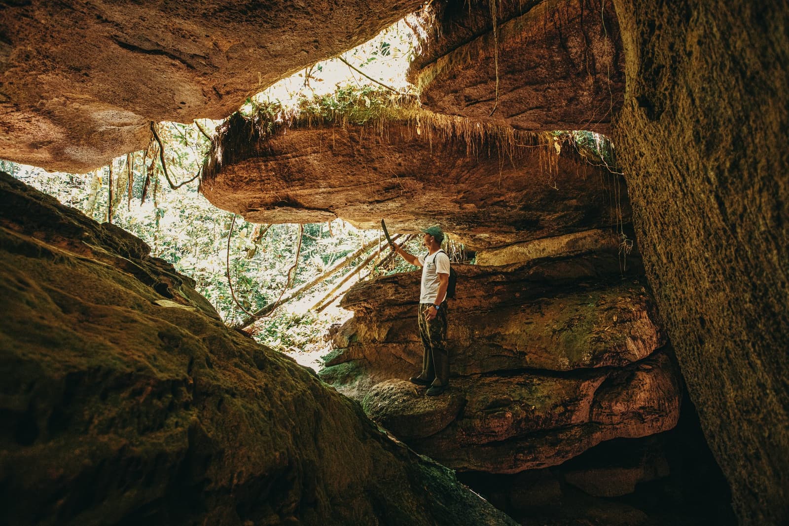 Some rooms took millions of years to build, and no reservation required. A guide stands inside the ancient sandstone cave formations of the Madadá Caves on a hiking excursion from Anavilhanas Jungle Lodge Brazil, holding a stick and gazing up at the dramatic overhanging rock ceiling draped with hanging roots