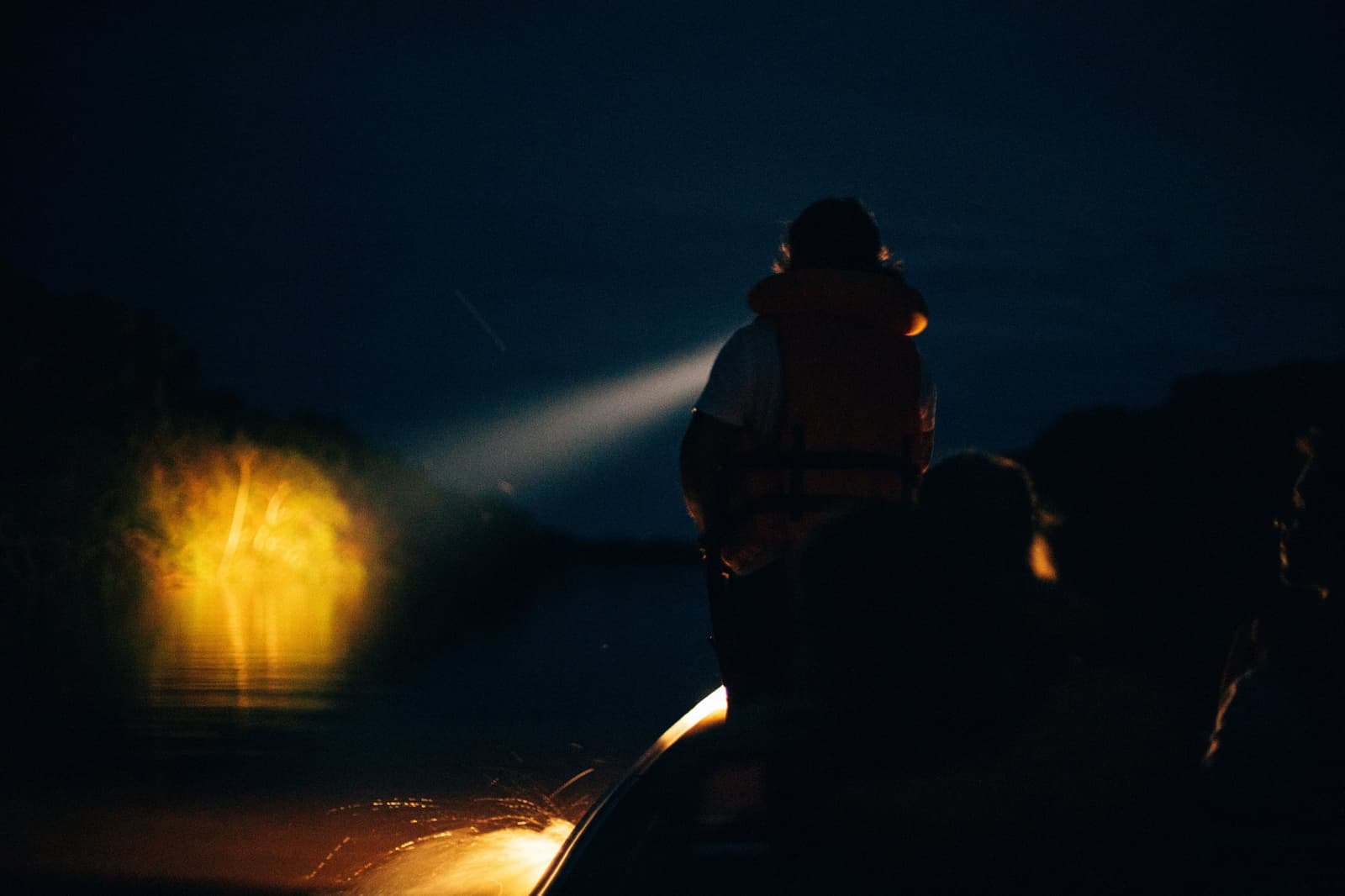 The river keeps its secrets close. The spotlight decides who gets a peek. A silhouetted guide in a life vest sweeps a spotlight across dark river water during a night tour at Anavilhanas Jungle Lodge Brazil, with a warm yellow lamp glowing at the bow
