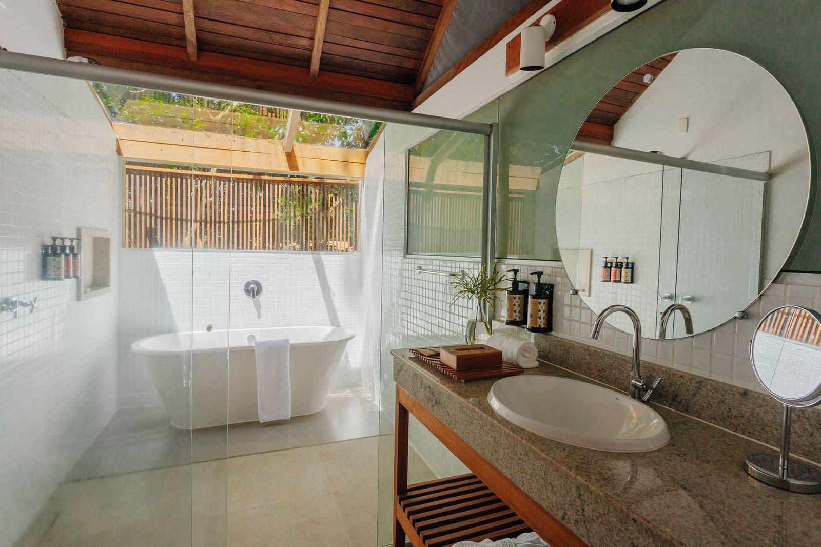 A bath with a forest view, because some comforts should come with canopy. Open-air bathroom at Anavilhanas Jungle Lodge Brazil featuring a freestanding soaking tub beneath a slatted wooden screen, granite vanity with round mirror, and a timber-beamed ceiling open to the treetops