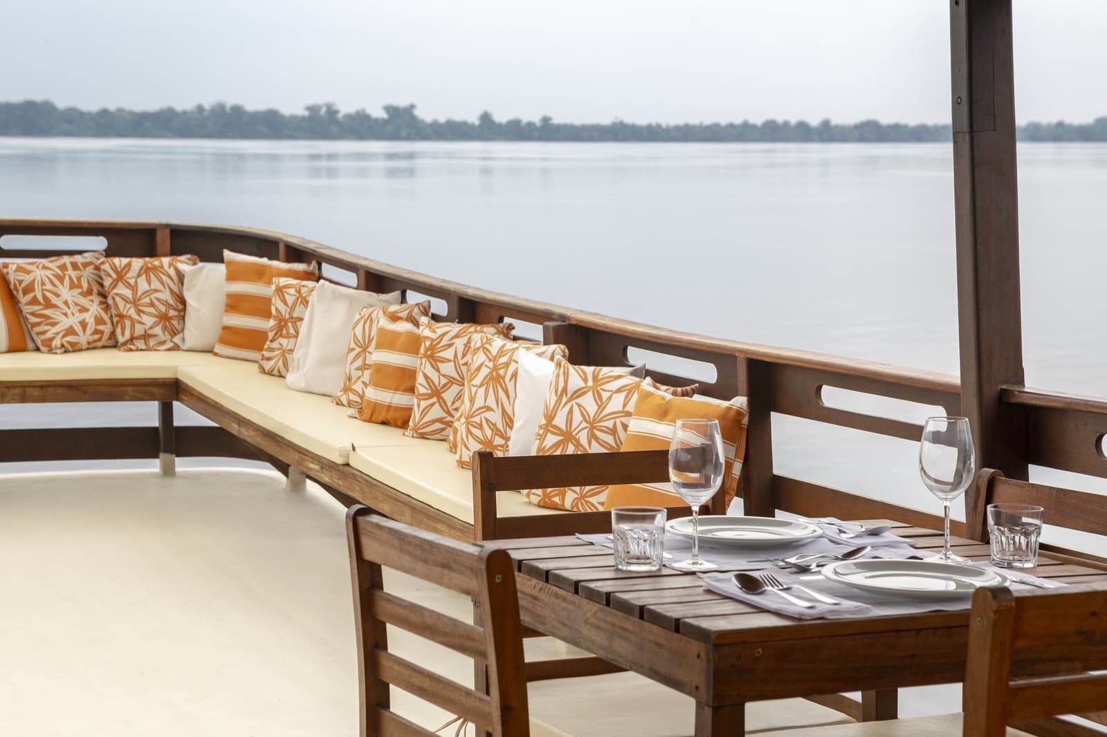 Dinner on the river, with the forest just close enough to feel it. A set dining table with wine glasses and cutlery on the open deck of a boat at Anavilhanas Jungle Lodge Brazil, with a cushioned lounge bench and a wide river stretching to the forested horizon