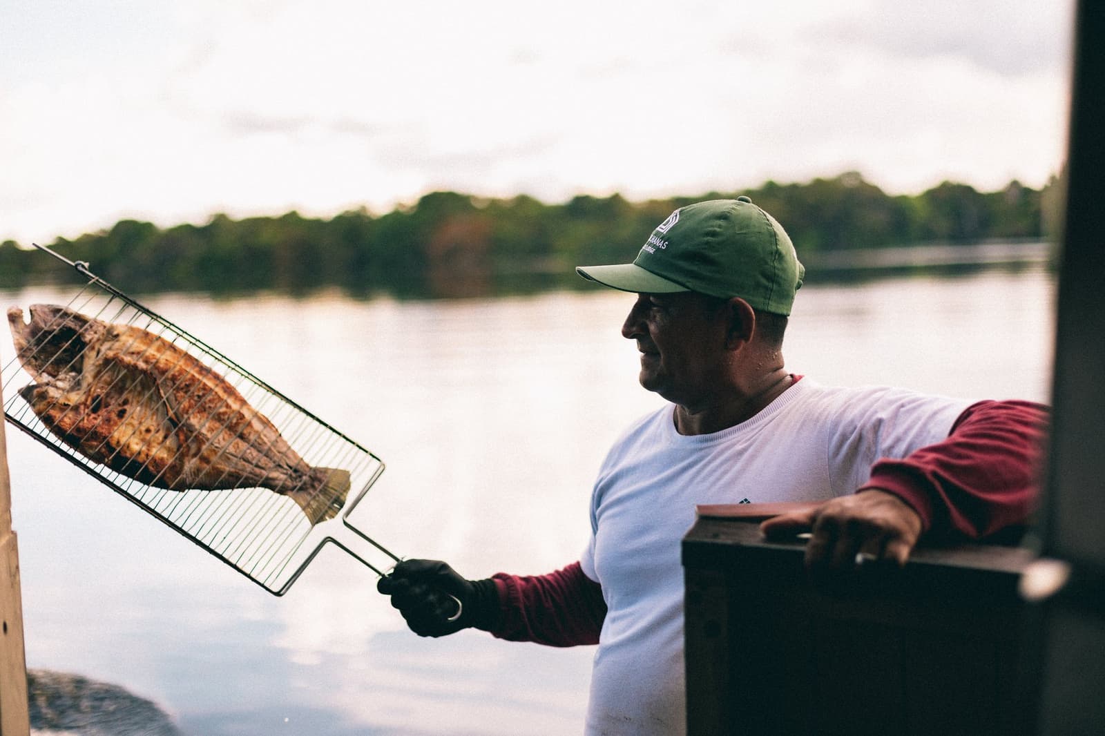 Fresh from the river, straight to the grill. Local cooking doesn't get more honest than this. A smiling man in a green cap holds a whole grilled fish in a wire basket over the Rio Negro at Anavilhanas Jungle Lodge Brazil, dense rainforest reflected in the calm water behind him