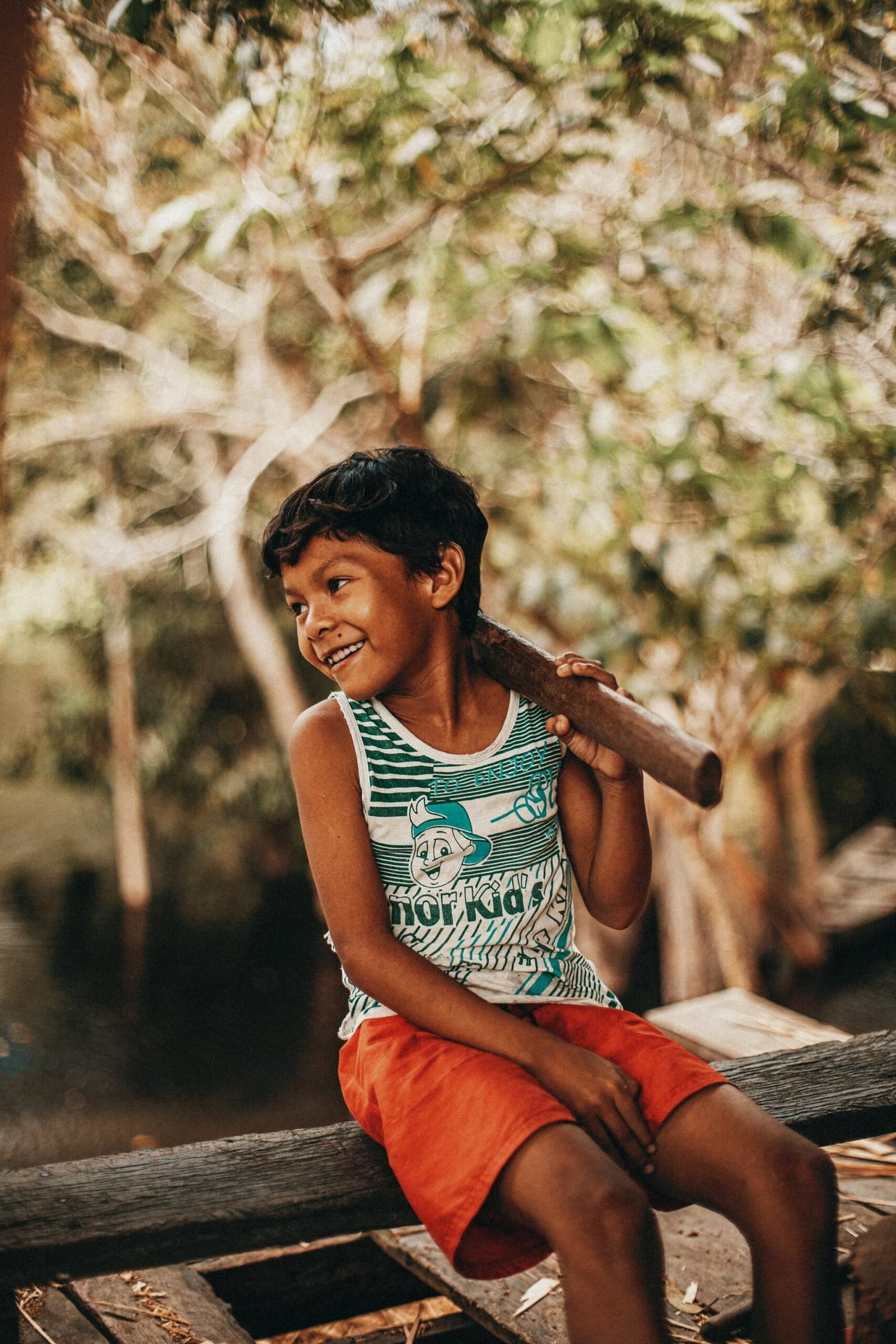 Some kids need no Wi-Fi to have the best afternoon of the week. A smiling boy sits on a wooden ledge holding a stick over his shoulder during a riverside community visit at Anavilhanas Jungle Lodge Brazil, with sun-dappled trees behind him