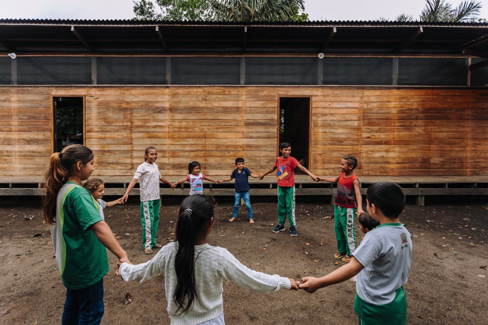 A circle of kids, a dirt yard, and enough joy to fill the whole forest. Children holding hands in a circle and playing outside a wooden community building during a riverside village visit at Anavilhanas Jungle Lodge Brazil
