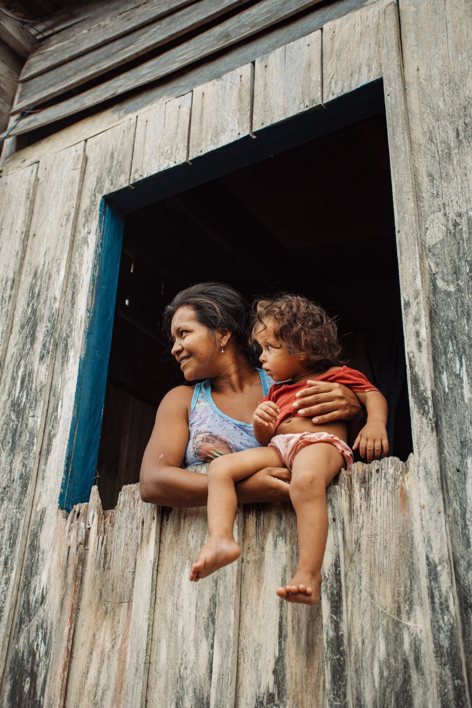 Life in the Amazon flows through its people, not just its rivers. A smiling woman holds a barefoot toddler on the sill of a weathered wooden window during a riverside community visit near Anavilhanas Jungle Lodge Brazil