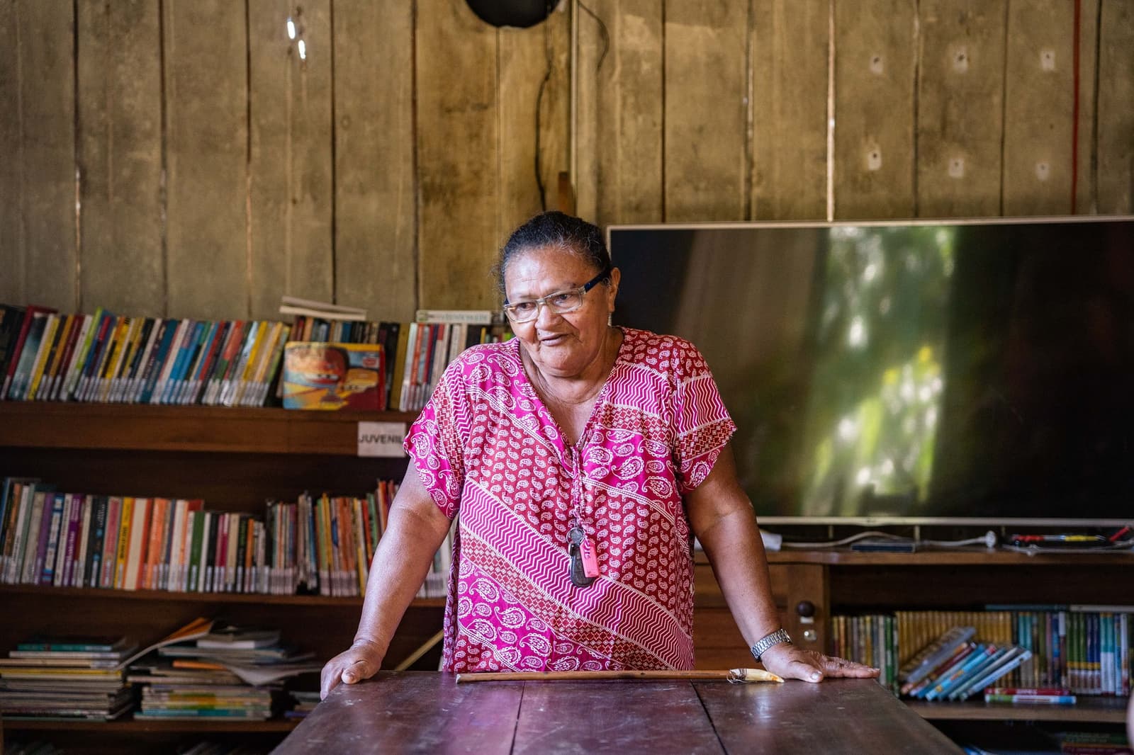 The kind of person who knows every book on those shelves, and every kid who borrowed them. A local woman in a pink patterned dress stands at a wooden table inside a community library near Anavilhanas Jungle Lodge Brazil, with colourful bookshelves lining the timber walls behind her