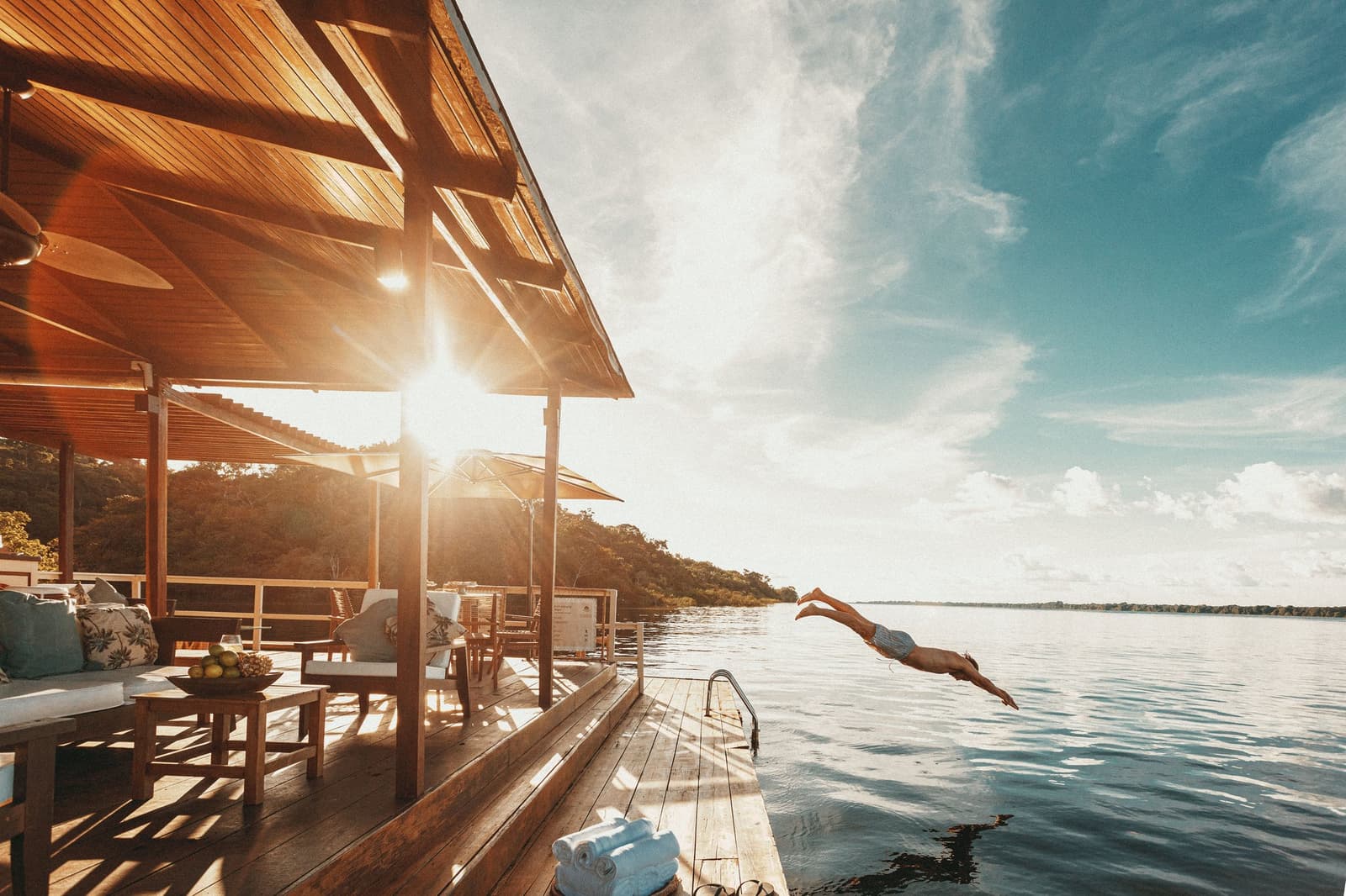 Some decisions are easy. The river is right there. A guest dives from the wooden deck of Anavilhanas Jungle Lodge Brazil into the calm Rio Negro at golden hour, with a furnished open-air lounge and lush forest visible in the background