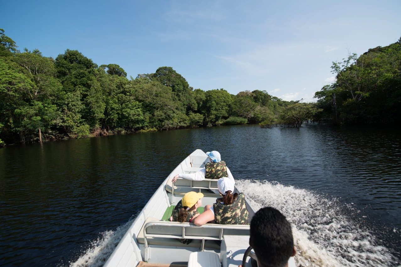 The forest closes in on both sides, and the only agenda is what's around the next bend. Guests wearing life jackets speed along a dark-water Amazonian river on a motorboat excursion at Anavilhanas Jungle Lodge Brazil, with dense rainforest canopy lining both banks under a clear blue sky