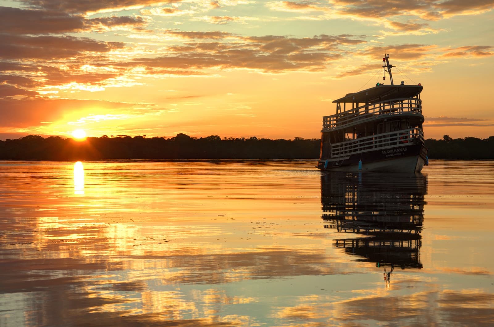 The river holds its breath at dusk, and the boat does the same. The Anavilhanas Jungle Lodge Brazil expedition boat moored on the glassy Rio Negro beneath a blazing orange sunset, its reflection perfectly mirrored on the still water