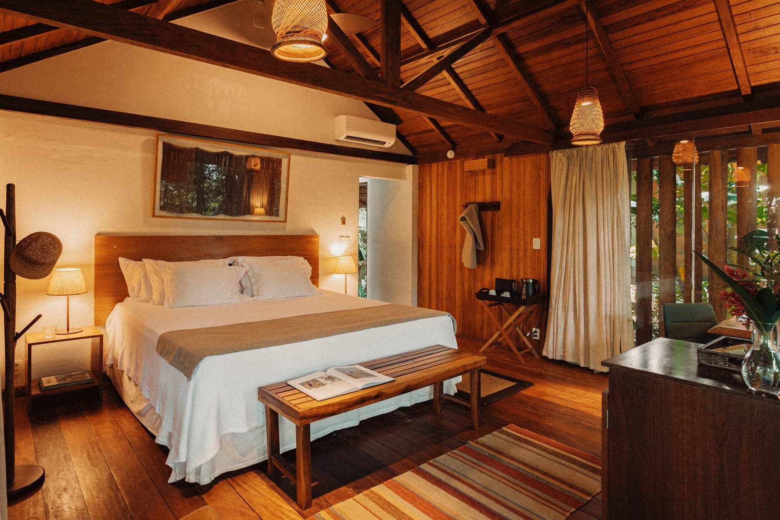 After a day on the river, this is exactly where you want to land. A warmly lit chalet bedroom at Anavilhanas Jungle Lodge Brazil, featuring a king-size bed with crisp white linens, exposed dark timber ceiling beams, woven rattan pendant lights, hardwood floors, and a window opening onto lush green foliage