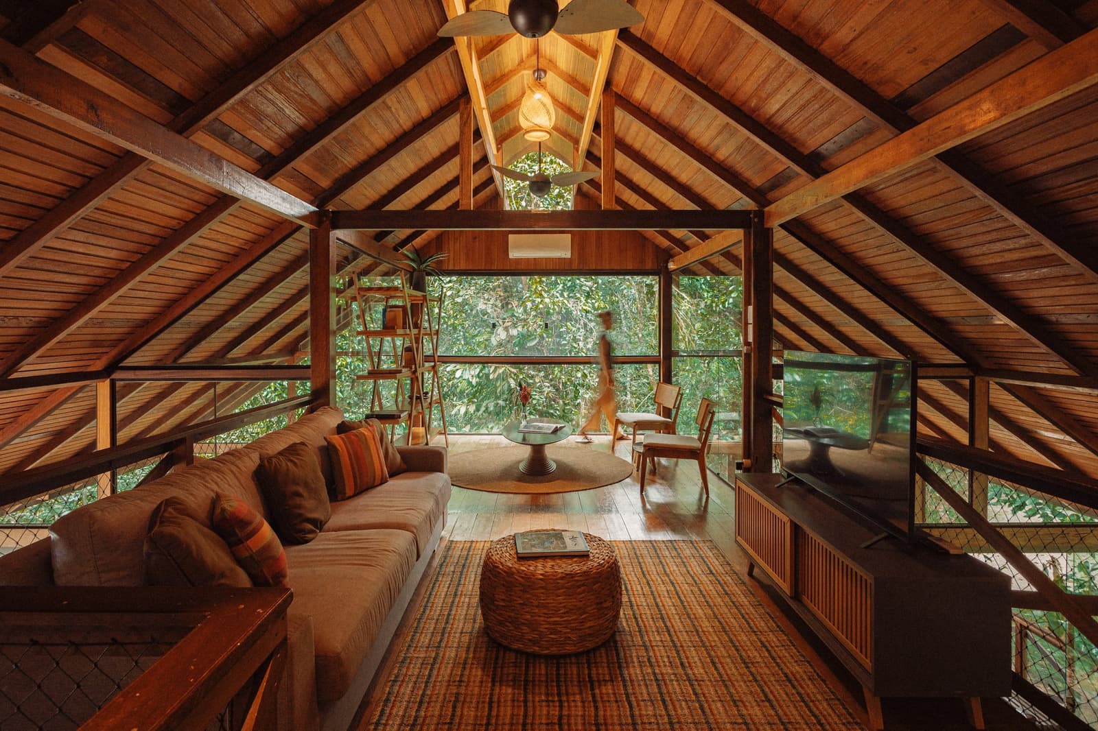 Your living room has a better view than most offices ever will. Vaulted timber ceiling and open-sided living room at Anavilhanas Jungle Lodge Brazil, with a plush sofa, woven rug, rattan ottoman and floor-to-ceiling views of dense rainforest canopy
