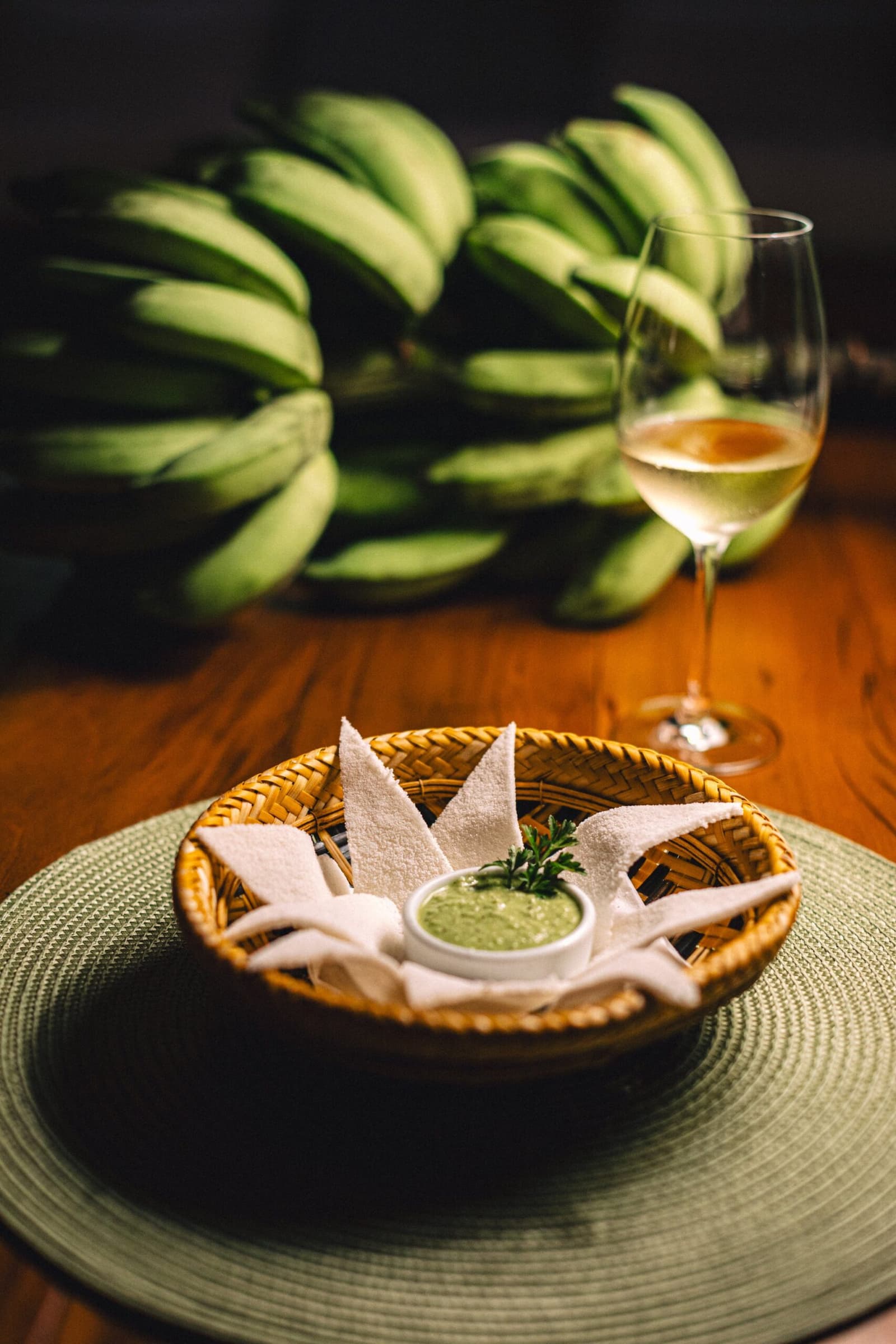 The Amazon's version of a warm welcome: simple, fresh, and hard to resist. A woven rattan basket holding white flatbread pieces arranged around a small bowl of green herb dip, served at Anavilhanas Jungle Lodge Brazil alongside a glass of white wine, with a bunch of green bananas softly blurred in the background