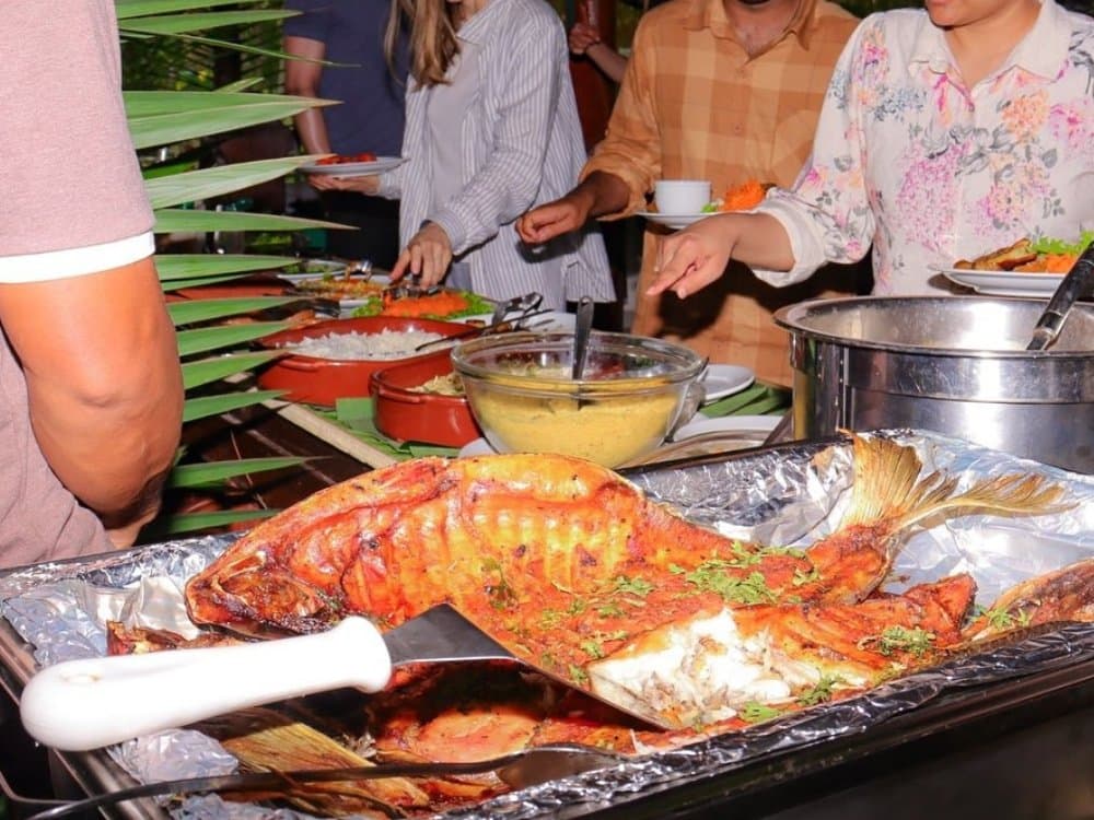 Fresh from the river — a whole roasted fish takes centre stage at the jungle buffet. A whole roasted fish garnished with herbs served on a foil tray at an outdoor buffet table on a brazilian amazon jungle tour, alongside rice, a golden sauce, and other dishes while guests help themselves