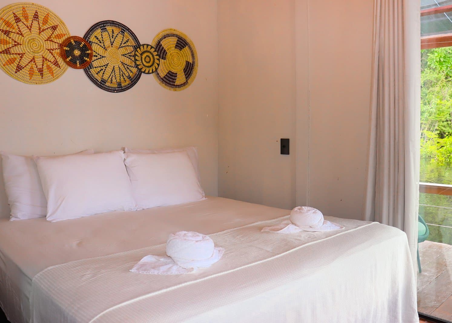 Handcrafted wall art, fresh linens, and the jungle just outside your window. A double bed with white linens and folded towels sits beneath woven basket wall art in a comfortable lodge room on a brazilian amazon jungle tour, with a window overlooking lush green forest