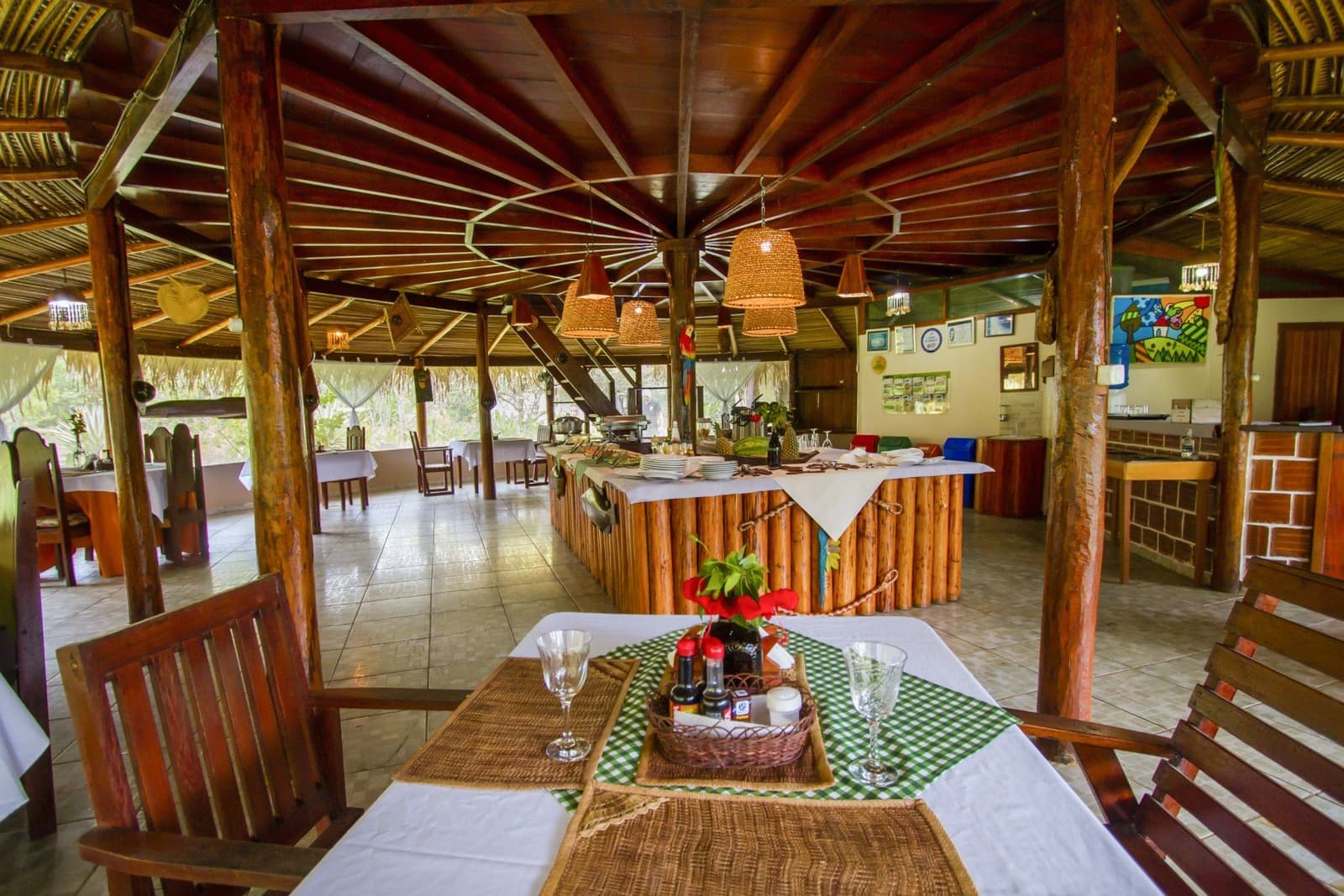 Rustic elegance under a thatched roof — meals served deep in the rainforest. Open-air thatched dining pavilion at an Amazon jungle lodge, with rustic log columns, wicker pendant lights, white-clothed tables, and a buffet counter set with plates and tropical fruit