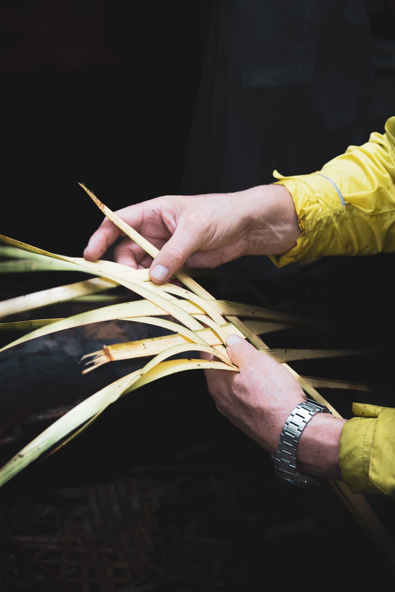 Nimble fingers, living fibers — jungle craft learned hands-on. Close-up of two hands in a yellow jacket weaving pale green palm leaf strips together during a brazilian amazon jungle tour survival skills lesson