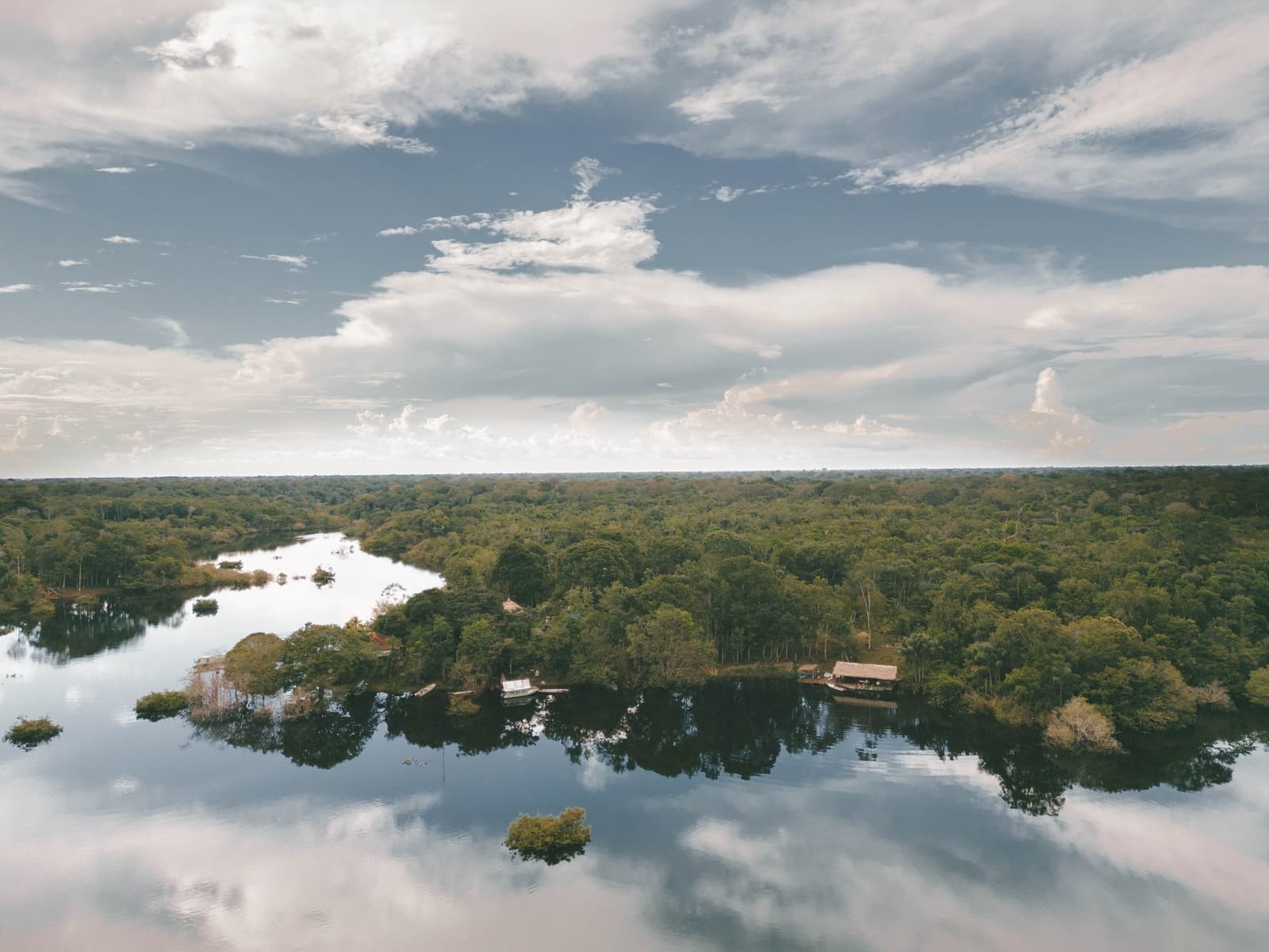 Canopy to the horizon, the lodge tucked quietly at the water's edge. Aerial view of a calm dark-water river winding through dense tropical rainforest on a brazilian amazon jungle tour, with thatched-roof lodge structures and a small boat visible along the tree-lined bank