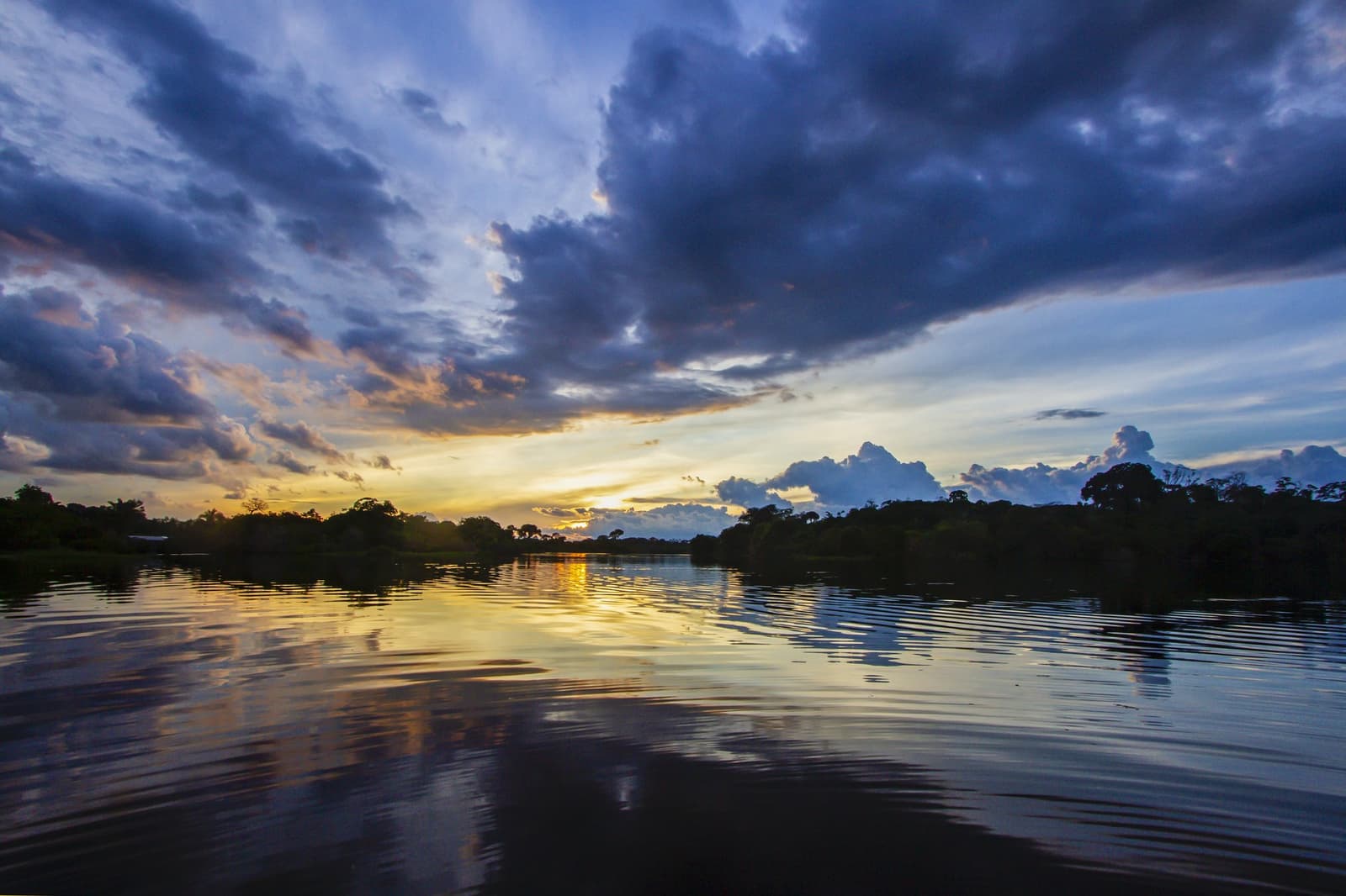 Storm clouds, golden water, jungle silence — dusk on the Amazon river. Photo: David Nascimento @ A dramatic sunset over a wide Amazonian river on a brazilian amazon jungle tour, with golden light reflecting off calm water and silhouetted rainforest trees lining both banks beneath deep blue storm clouds