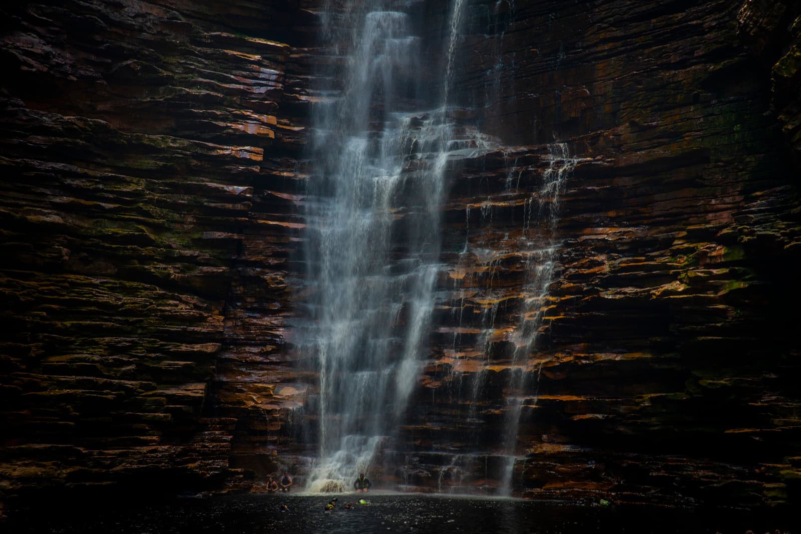 The kind of place that makes you forget what day it is. Tall multi-tiered waterfall cascading down layered sandstone cliffs on a chapada diamantina hike, two tiny figures visible at the base of the pool