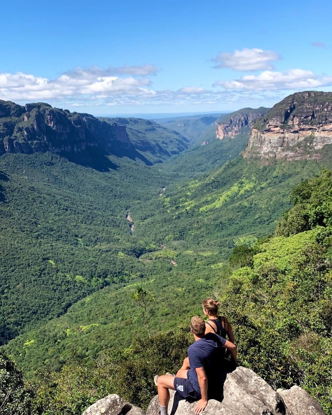 That moment when the valley does all the talking. Two hikers sitting on a rocky ledge overlooking a vast forest-filled canyon on a Chapada Diamantina hike, with towering sandstone cliffs and a river winding far below under a blue sky