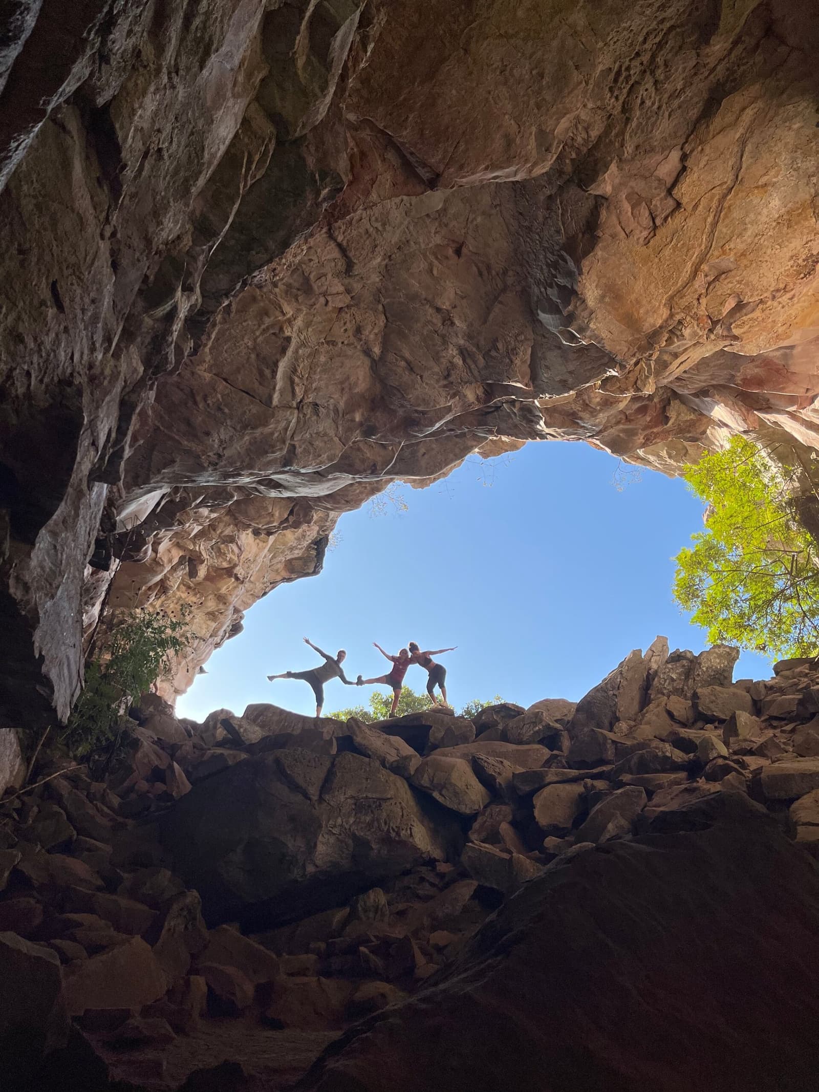 The cave sets the stage; they just brought the choreography. Two travelers strike yoga poses on boulders at the mouth of a large rock cave on a Chapada Diamantina hike, silhouetted against a bright blue sky and green tree canopy