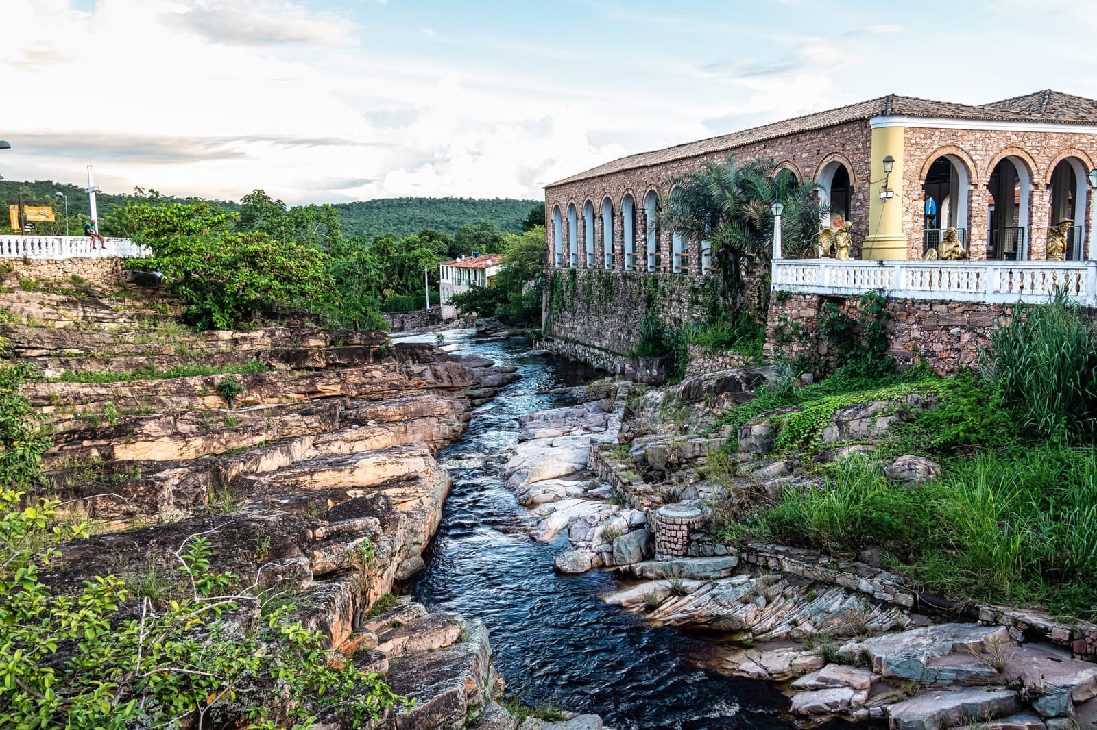 Town and river, sharing the same stone since forever. A historic stone building with arched colonnades overlooks a rocky river channel cutting through layered sandstone in a Chapada Diamantina hike base town, with forested hills rising in the background