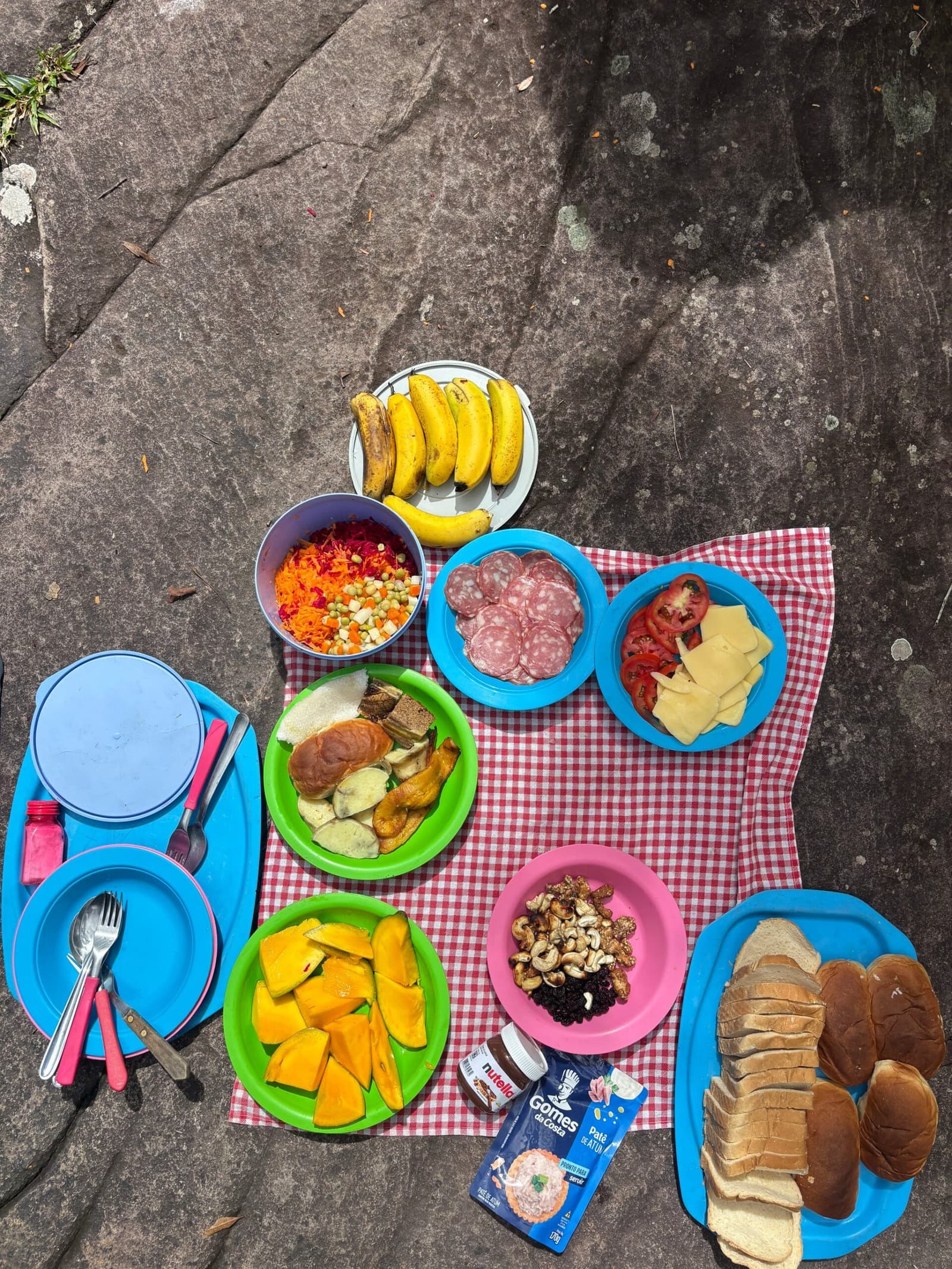 Lunch on a rock beats lunch at a desk, every single time. Overhead view of a colorful outdoor picnic spread on a flat rock during a chapada diamantina hike, with plates of sliced fruit, salami, cheese, tomatoes, grated vegetables, cashews, raisins, bananas, sandwiches, sliced bread, and Nutella on a red-checked cloth