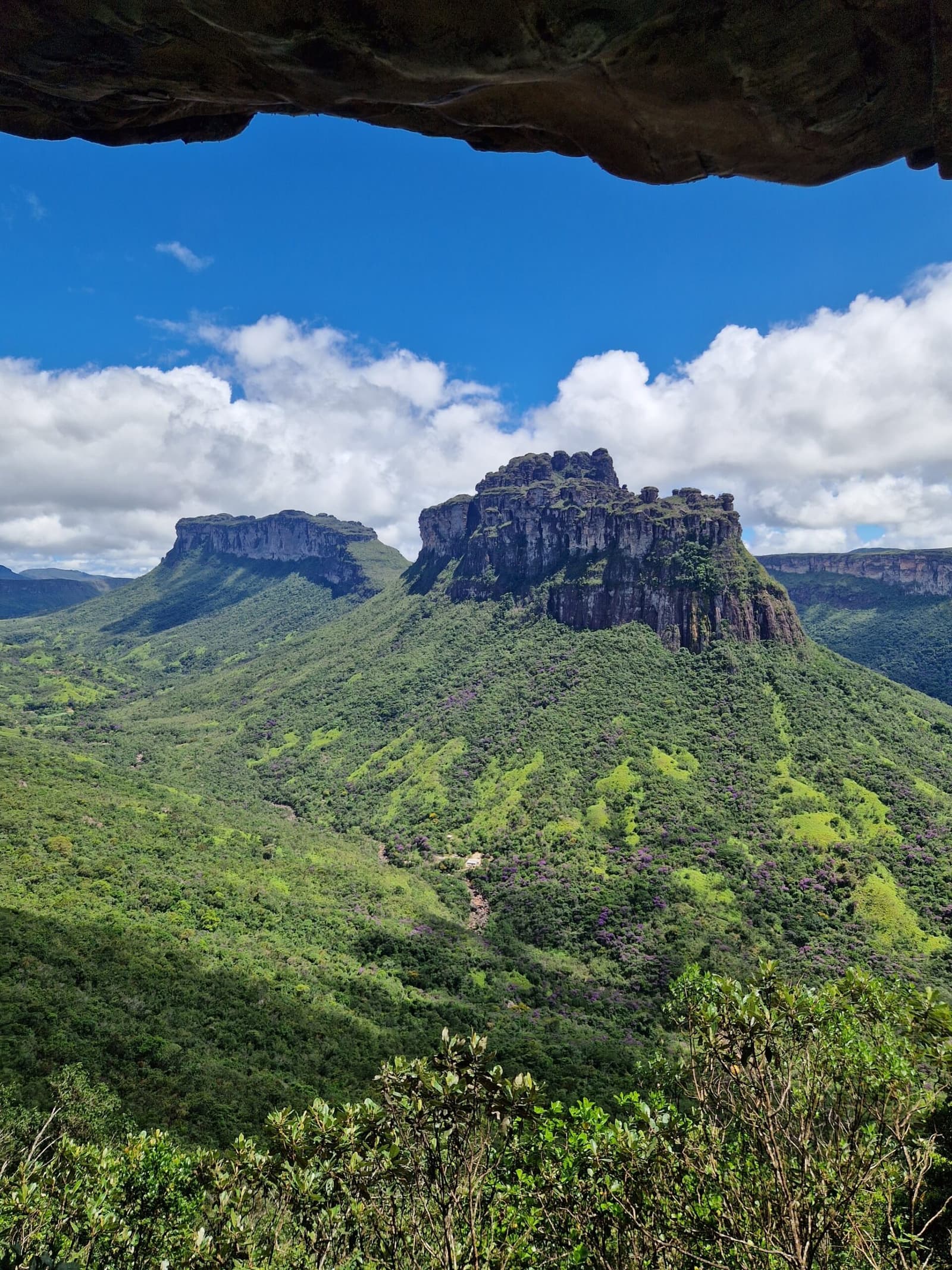 Nature built the frame; you just have to show up. A framed rock overhang frames a view of a dramatic flat-topped mesa rising above lush green forested hills on a chapada diamantina hike, under a vivid blue sky with billowing clouds