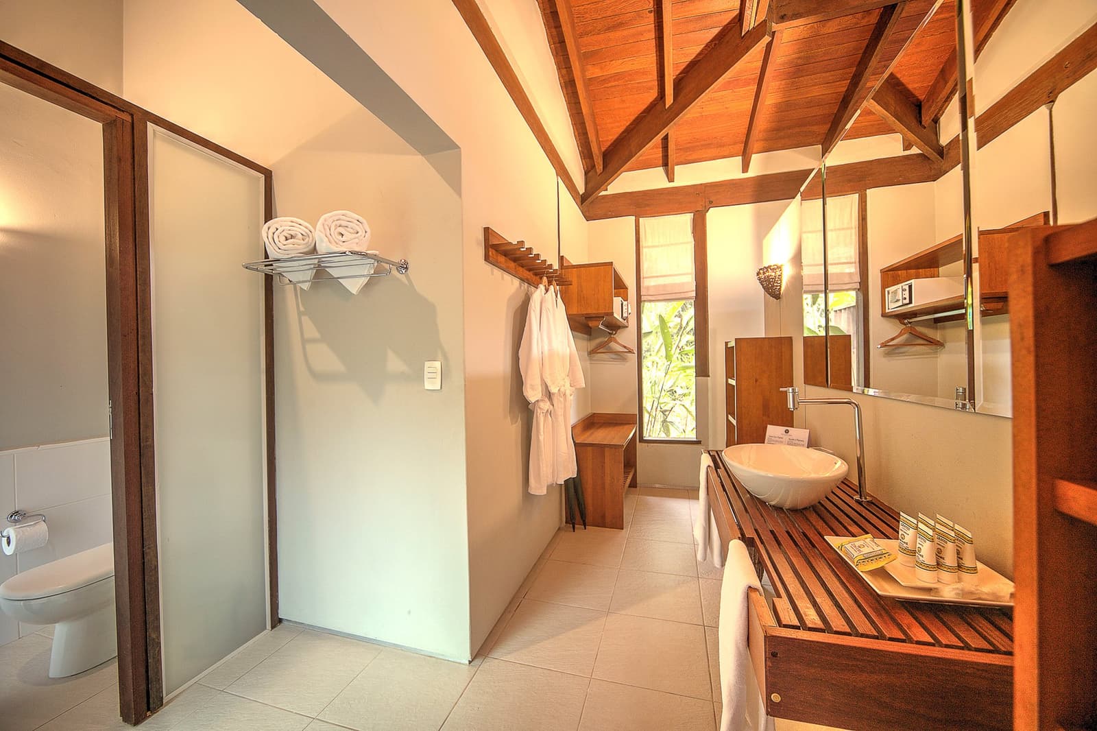 Comfort in the Amazon looks a lot like warm wood, fresh linens, and a window full of green. Spacious en-suite bathroom at Cristalino Jungle Lodge featuring a vessel sink on a slatted teak vanity, exposed timber roof beams, white bathrobes, rolled towels, and a glass-panel shower