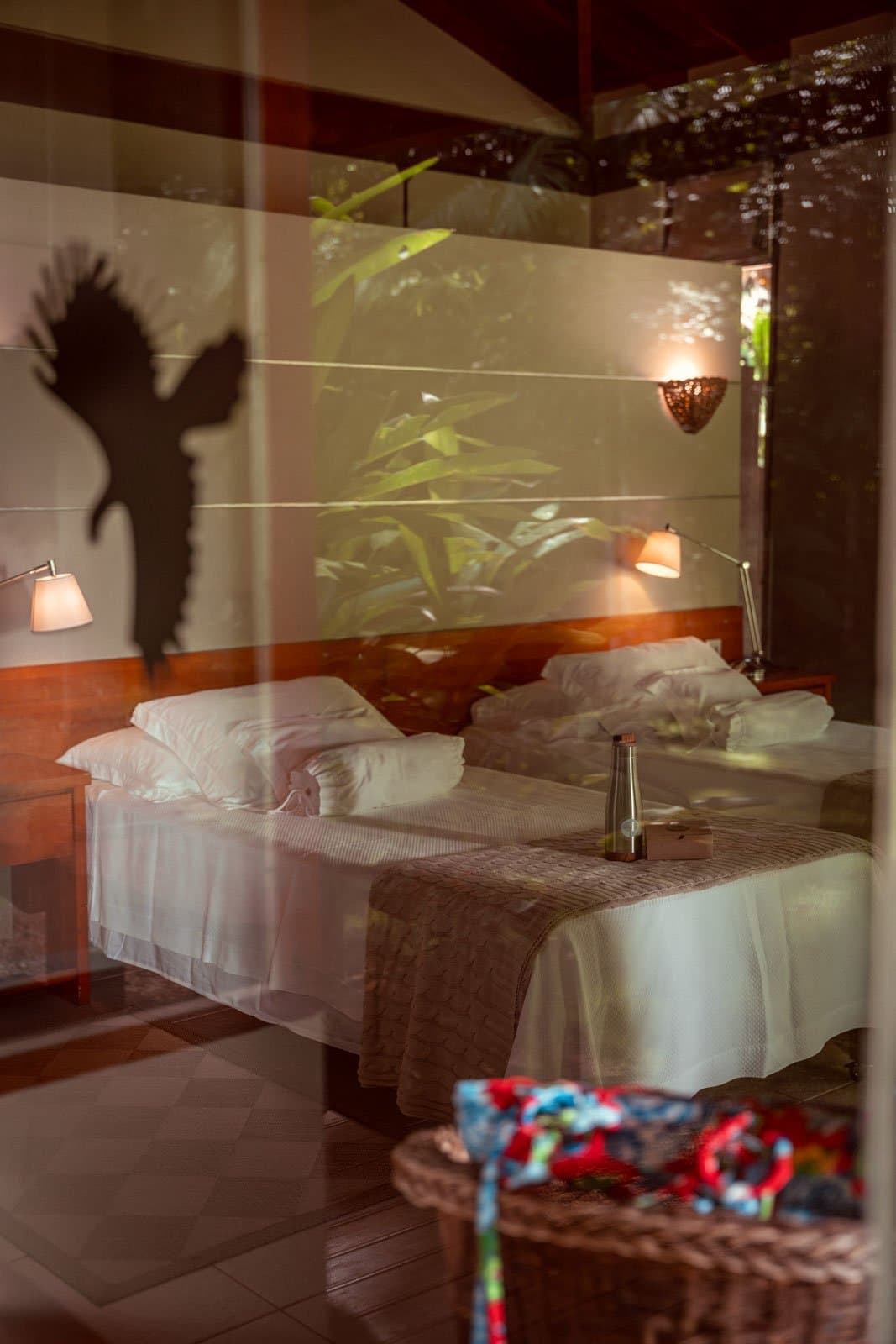Even the local wildlife finds a way into your room, sort of. A large bird silhouette cast on the wall of a Cristalino Jungle Lodge twin room, seen through a glass reflection, with warm bedside lamps and tropical foliage mirrored in the pane