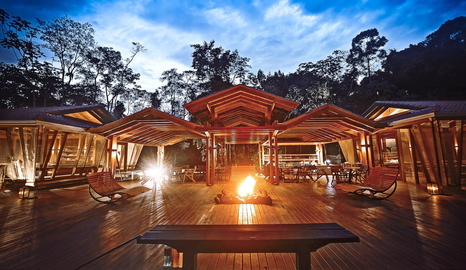 The fire pit earns its keep the moment the forest goes quiet. The open-air timber deck at Cristalino Jungle Lodge glows at dusk, with a crackling bonfire at its centre, rocking chairs on either side, a bar visible in the background, and tall rainforest trees silhouetted against a deep blue twilight sky