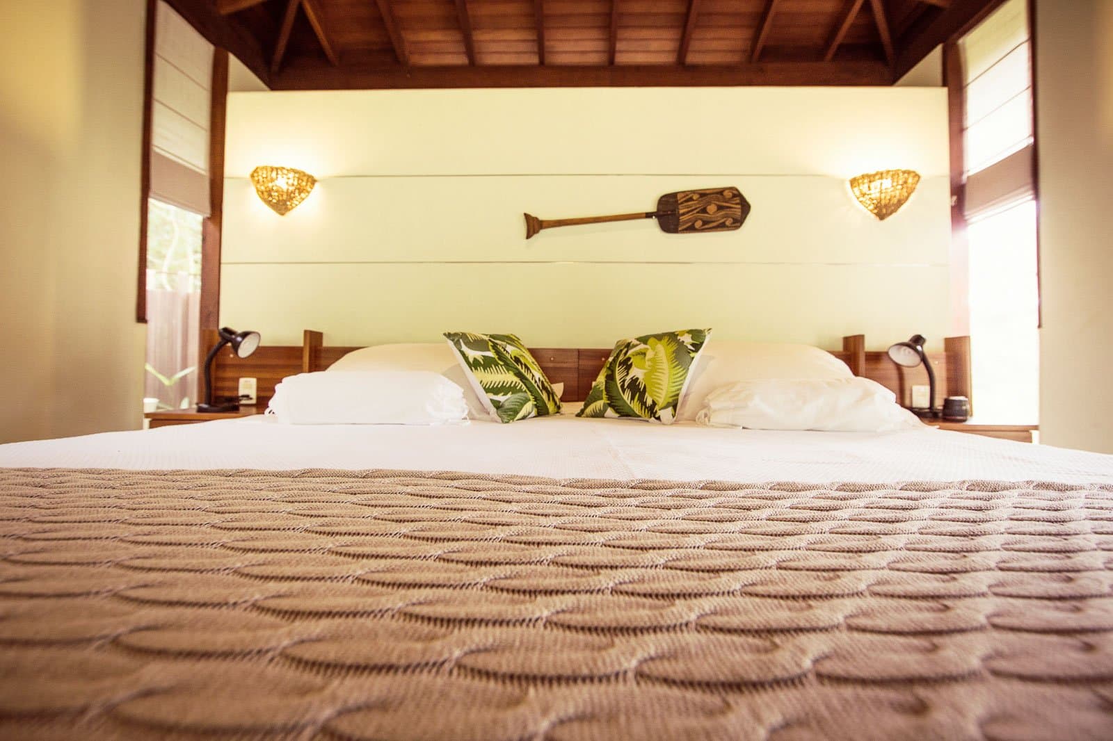 Crisp sheets, a wooden paddle on the wall, and the forest just outside the window. King-size bed with tropical leaf-print cushions, wooden headboard, woven wall sconces, and a carved paddle on the wall inside a Cristalino Jungle Lodge bungalow