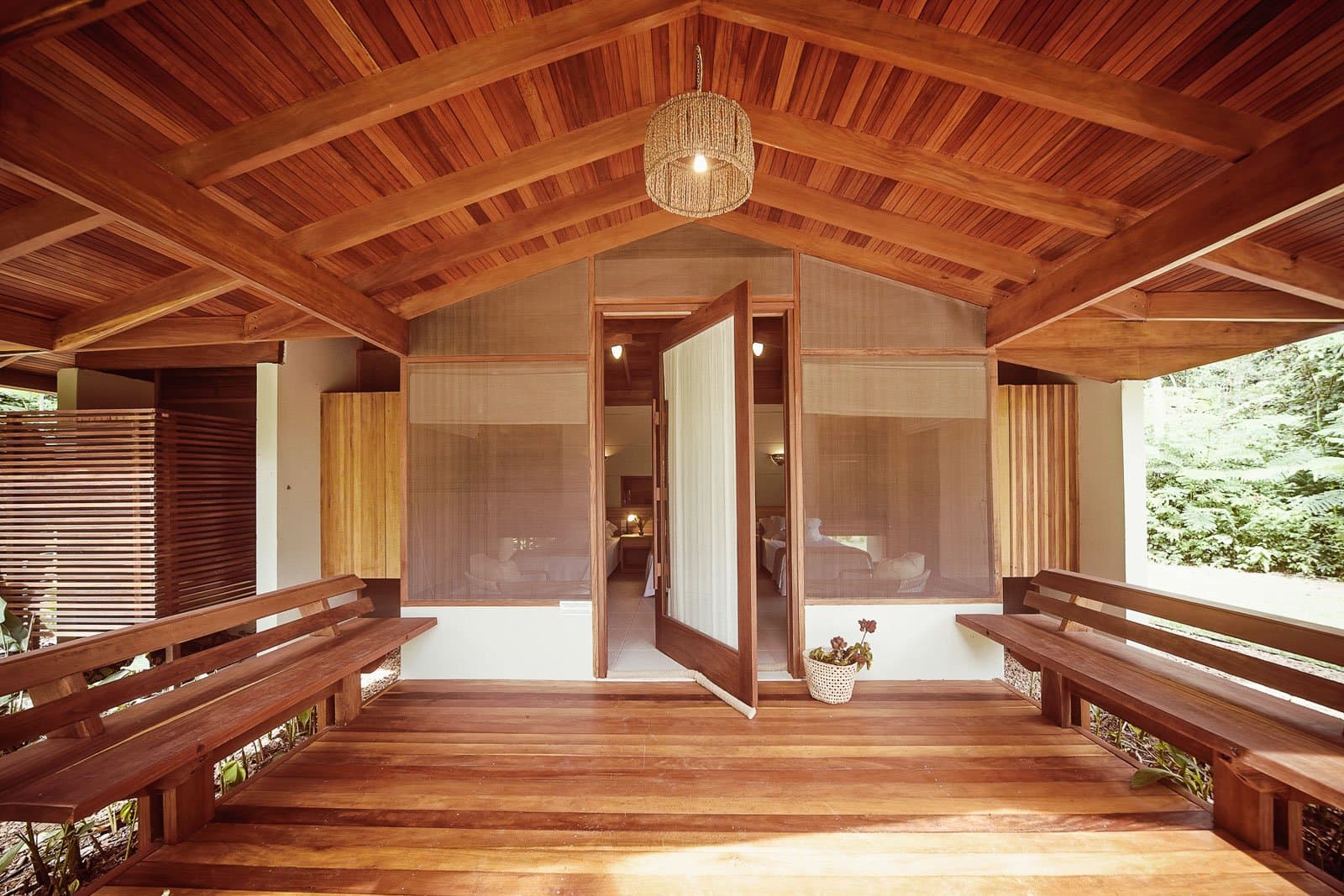 Come in, sit down, listen to the forest do its thing. A covered hardwood veranda at Cristalino Jungle Lodge with two built-in bench seats, a vaulted timber ceiling, woven pendant light, and open double doors leading into a bright guest bungalow surrounded by forest