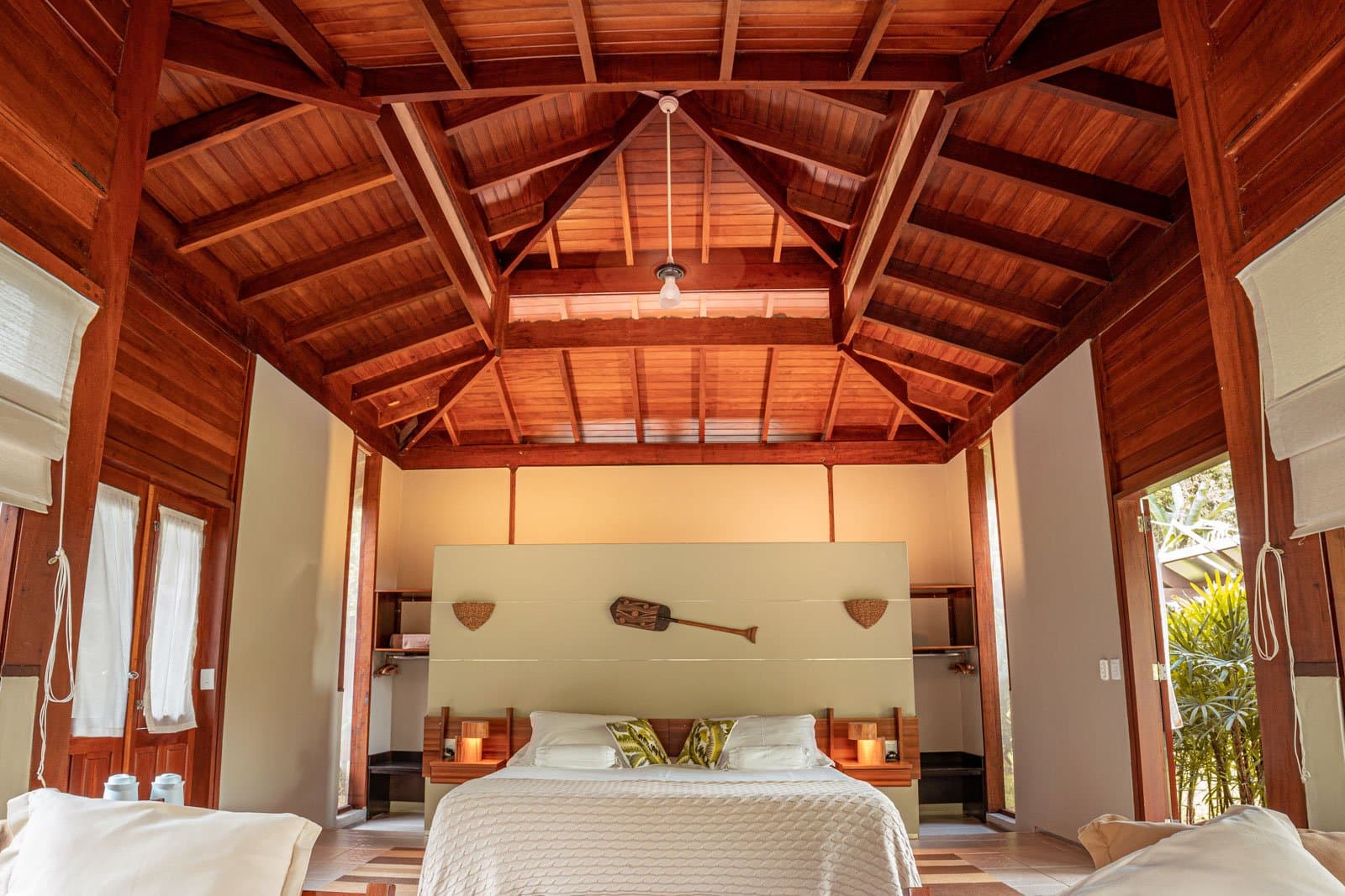 Sleep under soaring hardwood rafters, with a paddle on the wall to remind you the river is close. Spacious bungalow interior at Cristalino Jungle Lodge featuring a soaring vaulted hardwood ceiling, a king-size bed with white linens and leaf-print cushions, warm candlelit bedside lamps, and a decorative wooden paddle above the headboard