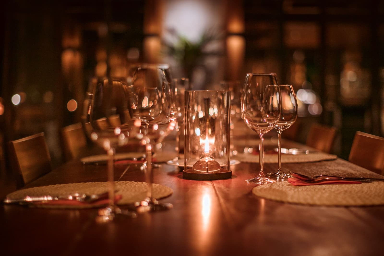 Good food tastes better when the forest is just outside the window. A long polished wooden dining table set for guests at Cristalino Jungle Lodge, with crystal wine glasses, woven placemats, red napkins, and a glowing glass candle lantern casting warm amber light across the room