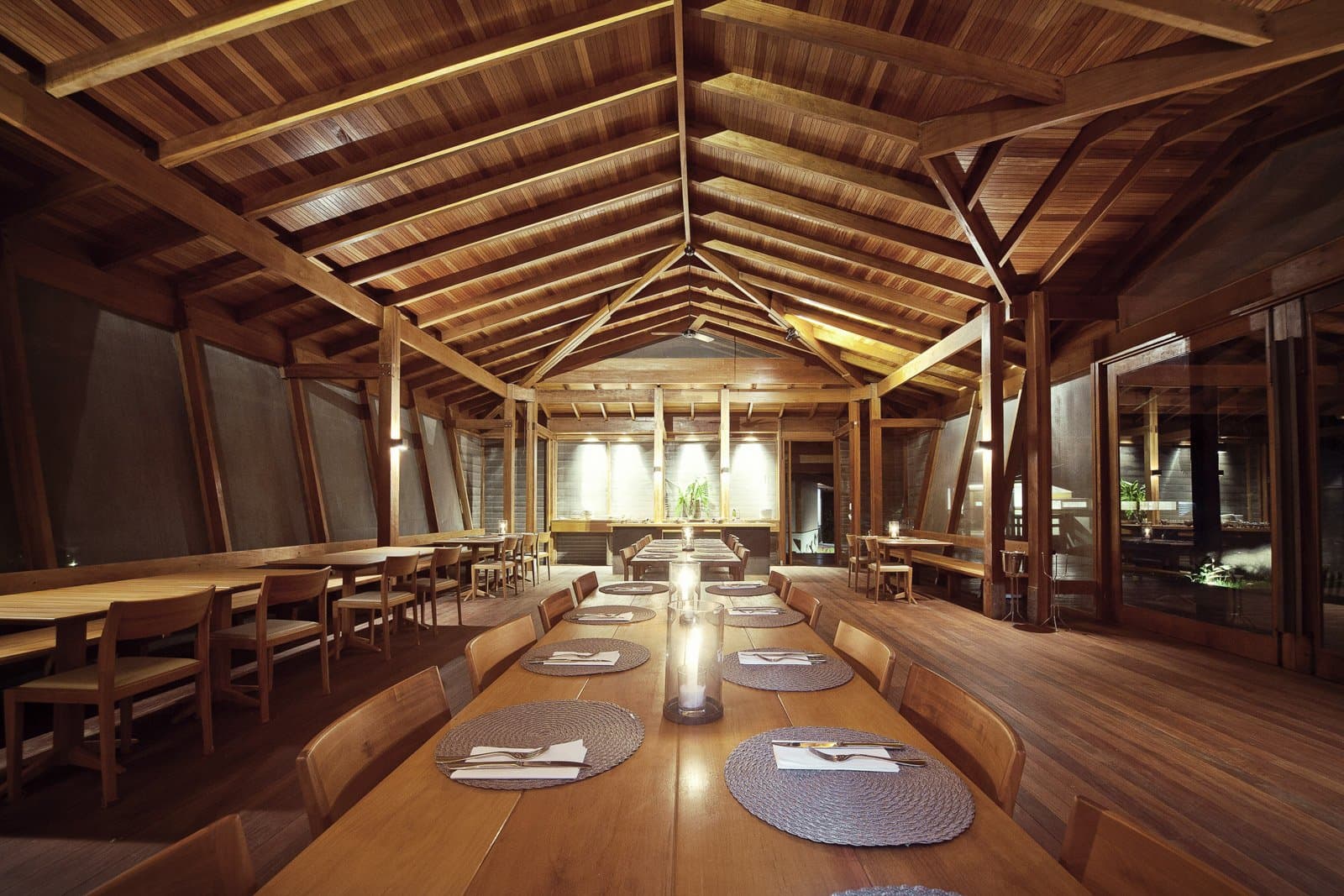Good food tastes better under a ceiling like this. The Cristalino Jungle Lodge restaurant dining room with a soaring vaulted timber ceiling, long communal wooden tables set with woven placemats and candles, and warm pendant lighting