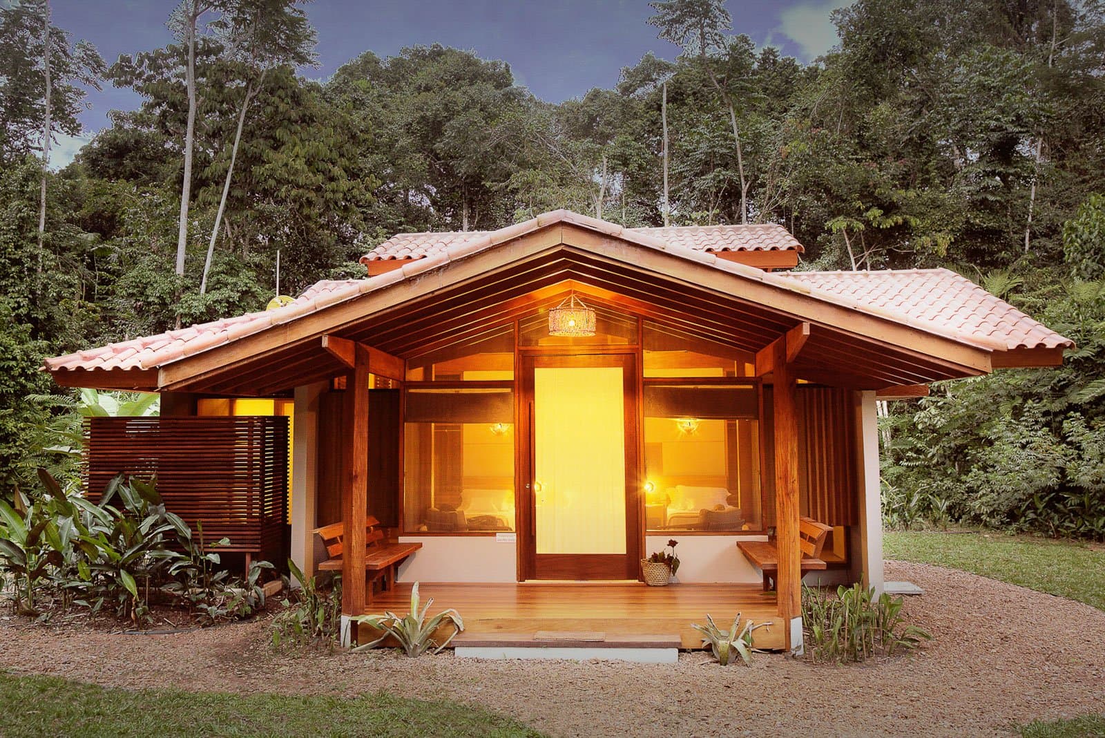 Your room for the night, with the forest doing all the decorating. A timber-framed bungalow at Cristalino Jungle Lodge glows with warm amber light at dusk, its terracotta-tiled roof and wooden deck framed by dense rainforest canopy