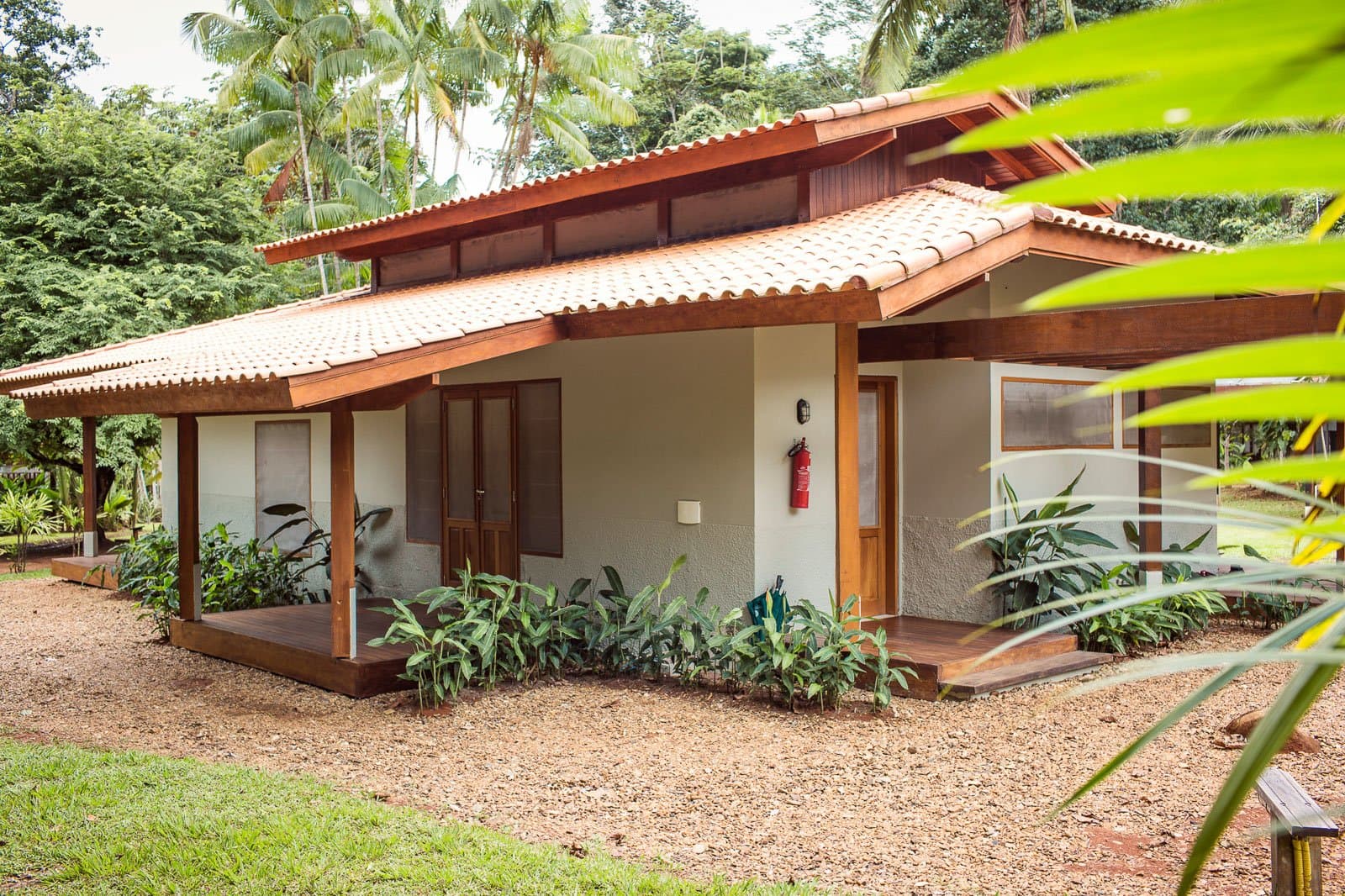 Your base for the Amazon, comfortable enough to forget you're deep in the jungle. Exterior view of a Cristalino Jungle Lodge chalet with a terracotta-tiled roof, rich hardwood trim, shaded veranda, and lush tropical planting set against a dense green forest backdrop