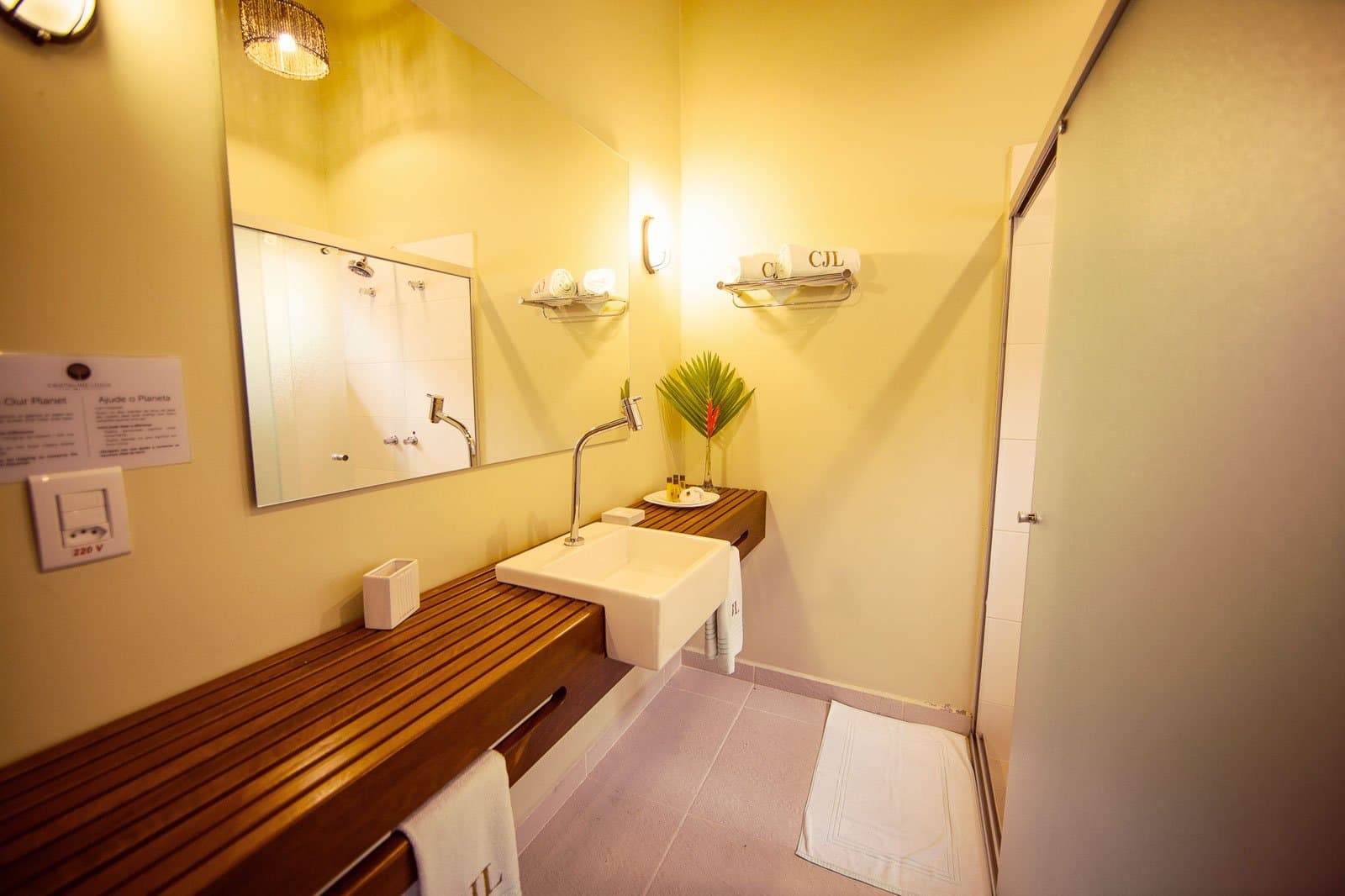 Comfort in the Amazon doesn't mean roughing it, this bathroom proves the point. Bright en-suite bathroom at Cristalino Jungle Lodge featuring a square vessel sink on a teak slatted vanity, warm wall sconces, monogrammed towels, and a red heliconia bloom beside the shower