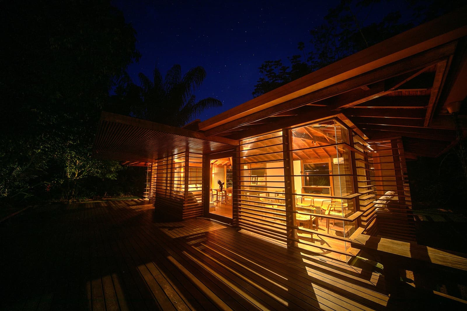Dinner hour in the forest, where the jungle goes quiet and the lodge lights up. Cristalino Jungle Lodge glowing from within at night, its timber-slatted walls and wide wooden deck illuminated by warm amber light beneath a star-filled sky
